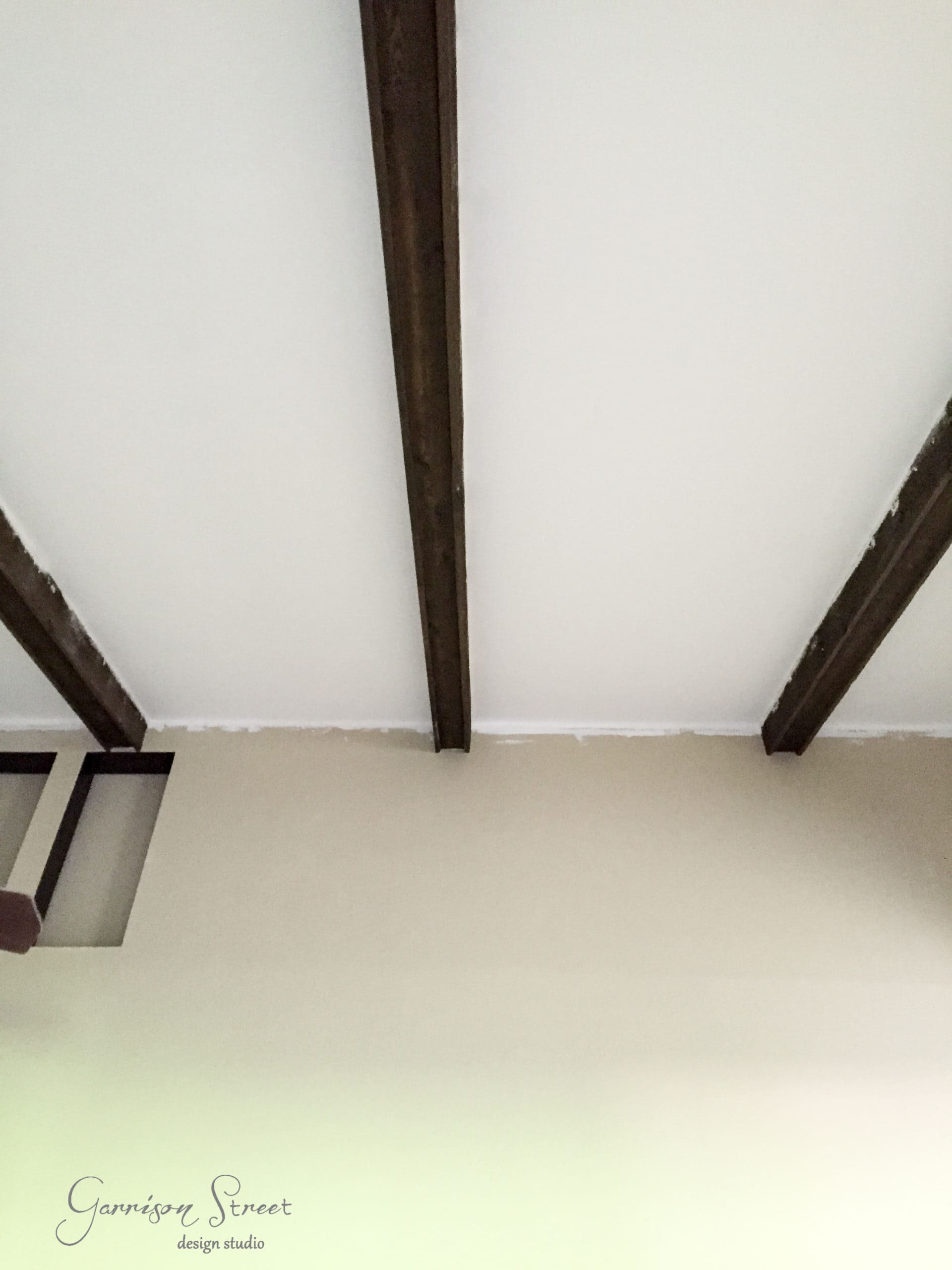 A ceiling featuring three exposed dark wooden beams against a white surface. A portion of a beige wall is visible at the bottom, and a modern light fixture is partially seen on the left. Garrison Street Design Studio logo is in the corner.