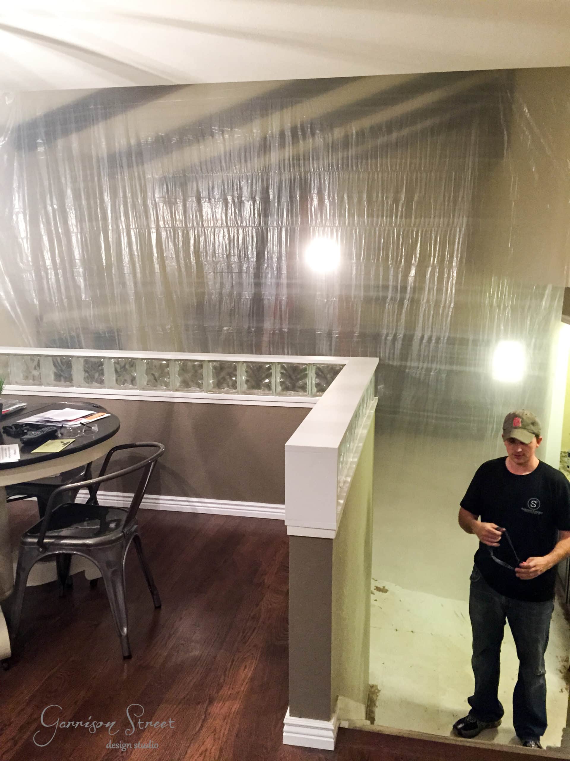 Man stands next to a staircase covered with plastic sheeting in a home under renovation. A small round dining table with chairs is visible nearby, with various papers on its surface. The environment suggests active construction or remodeling.