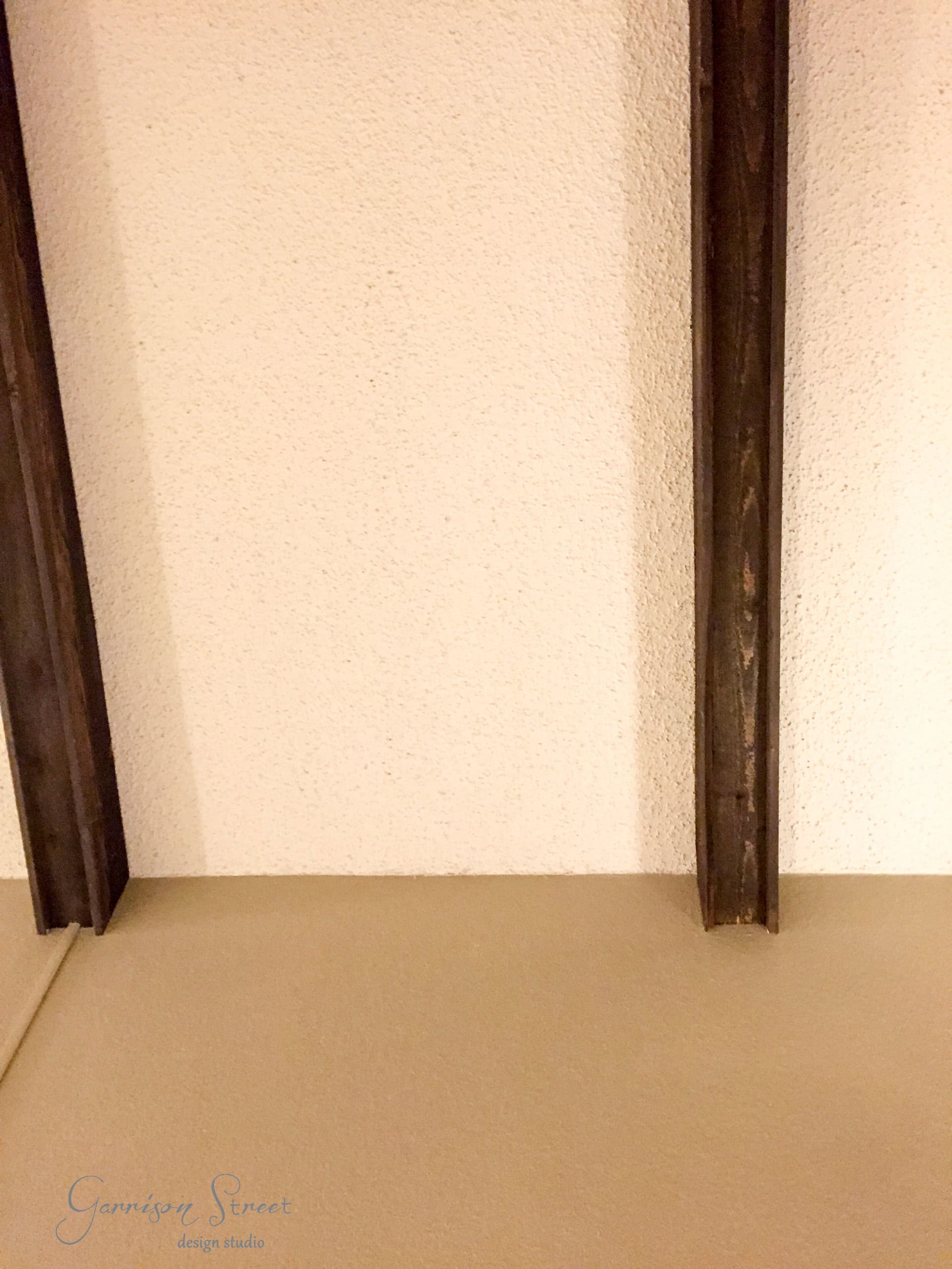 A textured white ceiling with three exposed dark wooden beams. The lower part of the image shows a beige wall and the corner where it meets the ceiling.