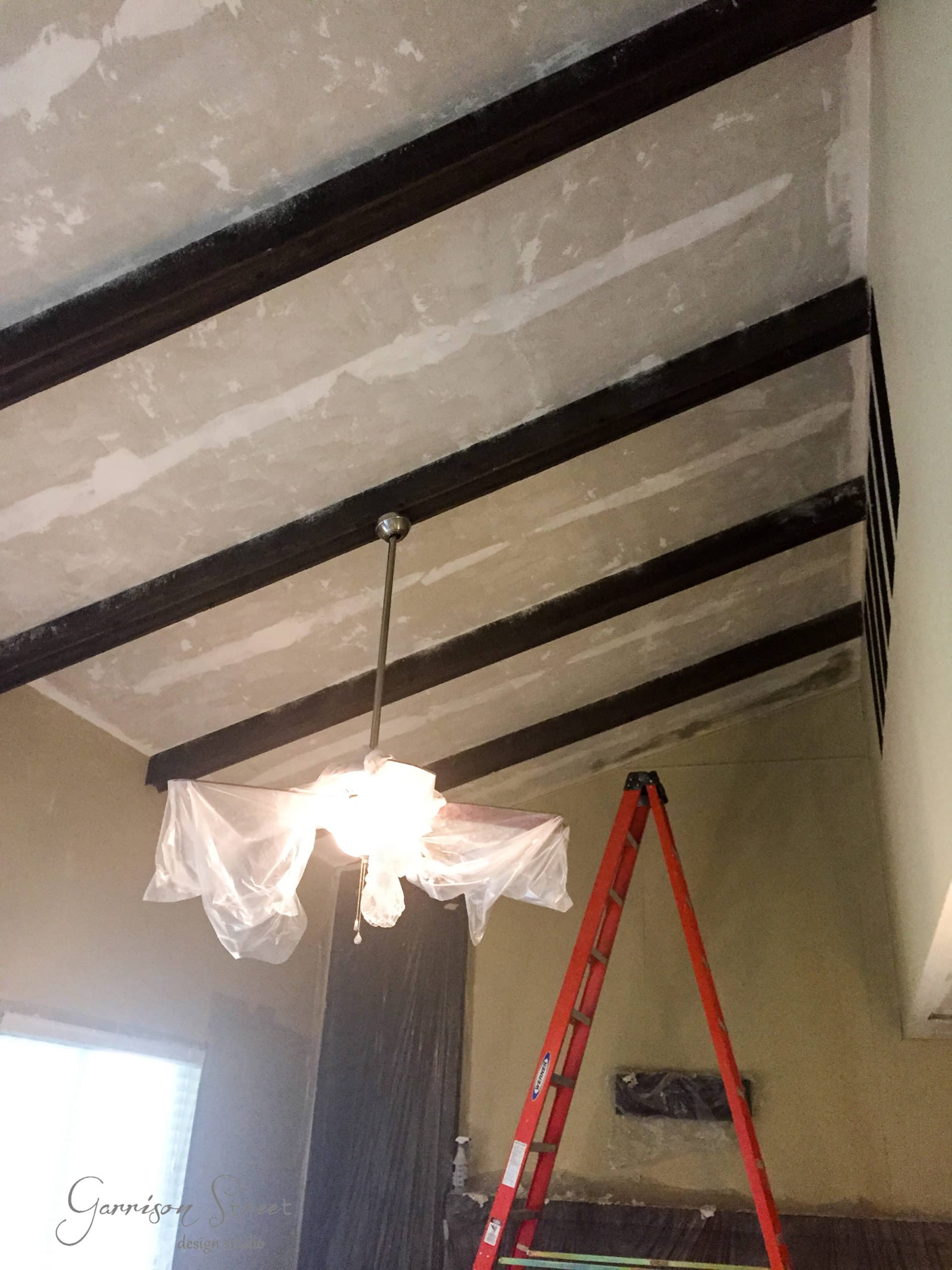 A room with a high, angled ceiling featuring exposed wooden beams. A red ladder is propped against a wall. The light fixture is covered with plastic, possibly for protection during renovation or painting work.