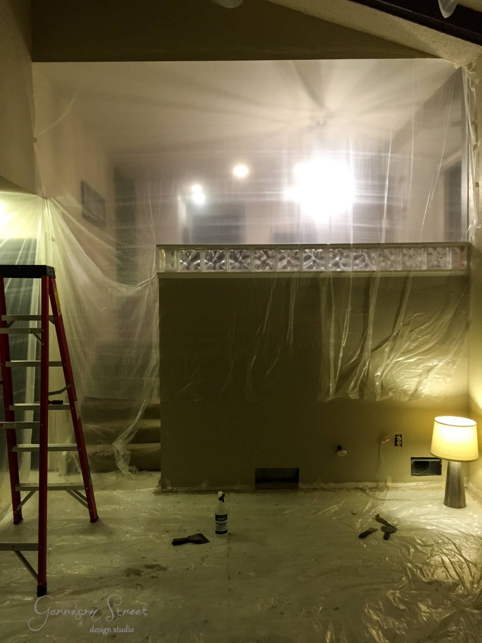 A room under renovation with plastic sheets covering walls and floors. A red ladder stands on the left, and a lit lamp is on the right. Tools and a paint can are visible on the floor. Ceiling lights shine brightly through the plastic.
