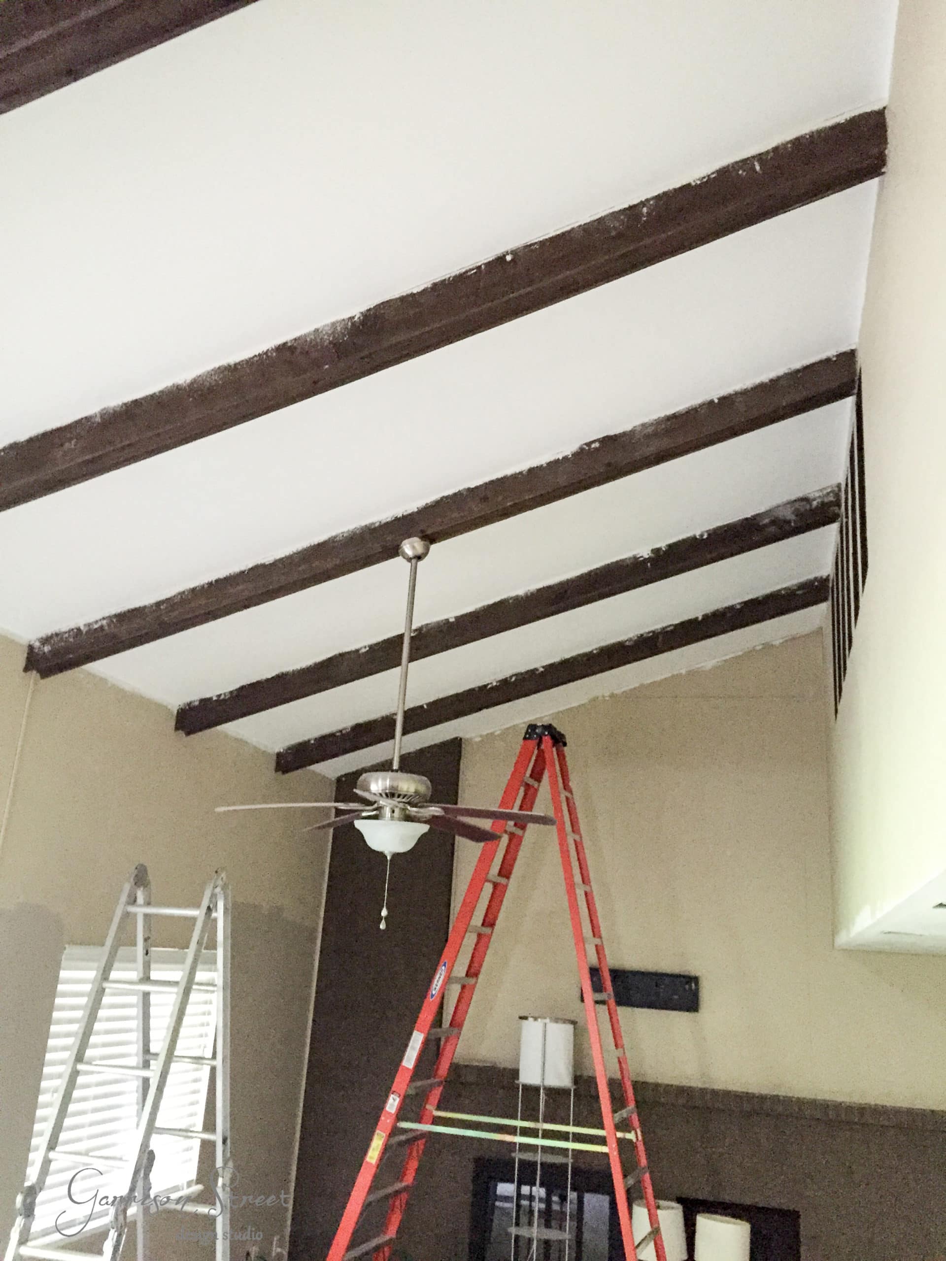 A room with a vaulted ceiling featuring exposed wooden beams. Two ladders are set up under a ceiling fan, suggesting maintenance or construction work. Light enters through a window on the left, illuminating the neutral-toned walls.