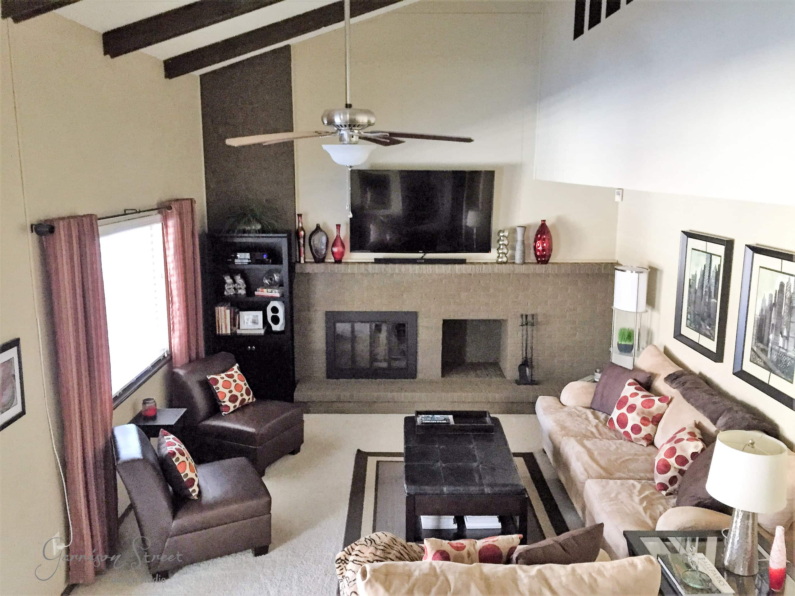 A cozy living room with a beige sectional sofa and two brown armchairs arranged around a dark coffee table. A fireplace with a mounted TV above is central, flanked by red vases. Large windows and decorative ceiling beams add to the room's charm.