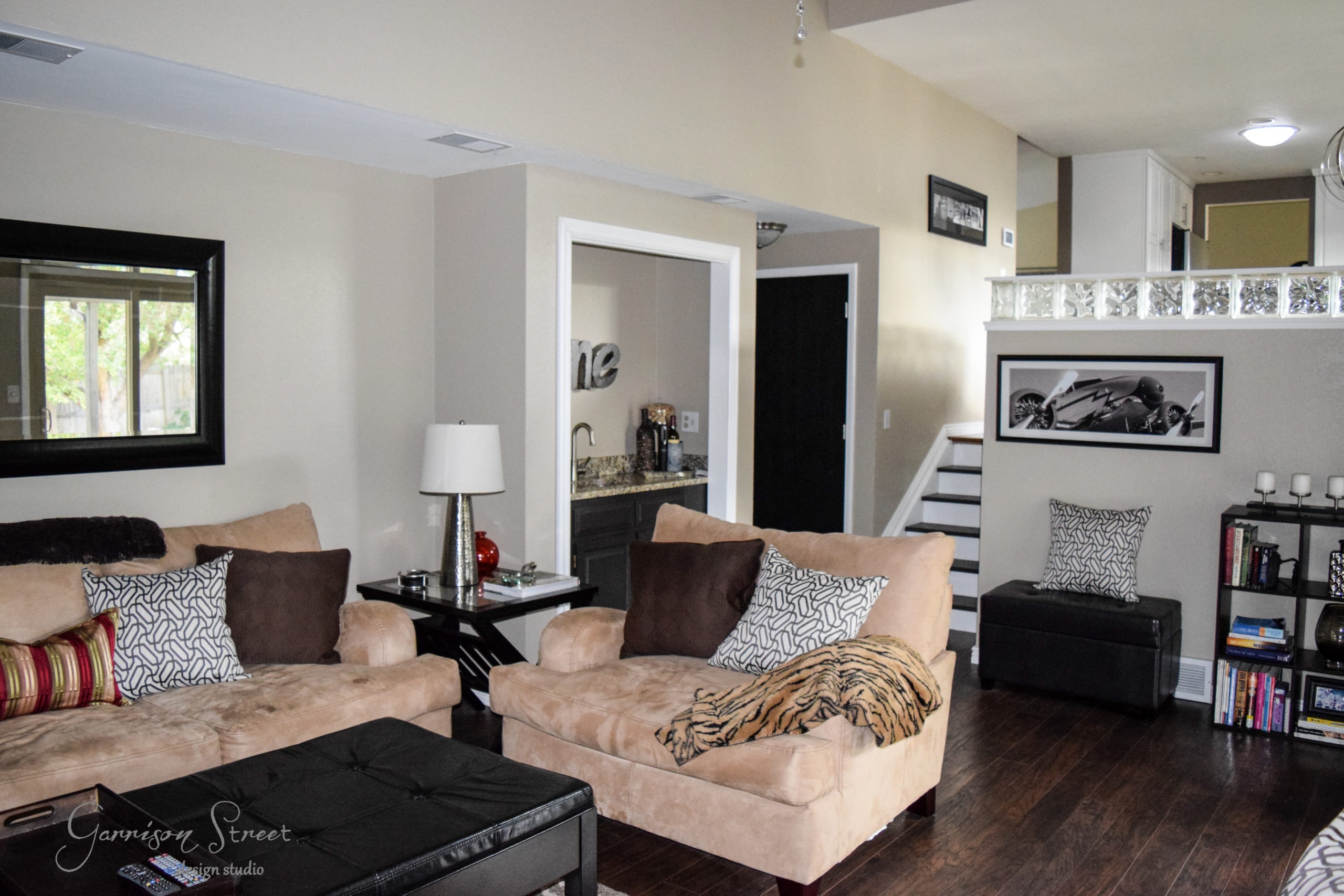 A cozy living room with beige sofas and patterned pillows. A small ottoman, wooden floor, and a patterned rug enhance the space. A staircase leads to an upper area, and a bookshelf with decor sits against the wall. A large window lets in natural light.