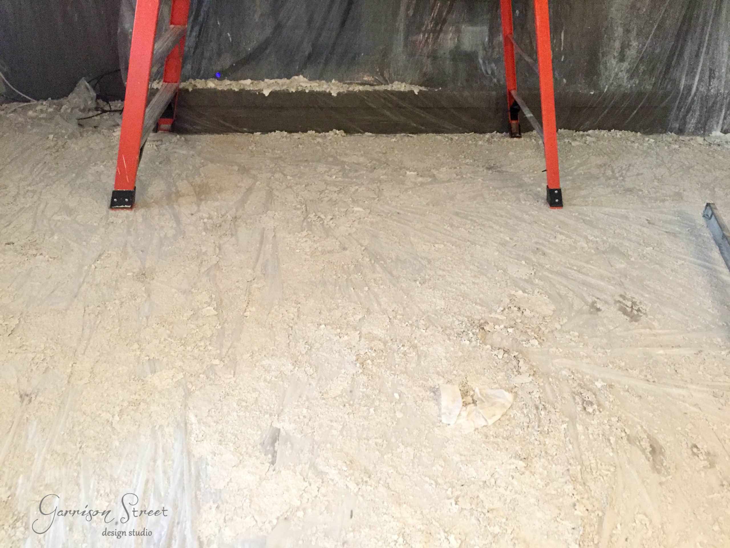 A construction site with a red ladder on a dusty, debris-covered floor. The background has a plastic sheet draped along the wall.