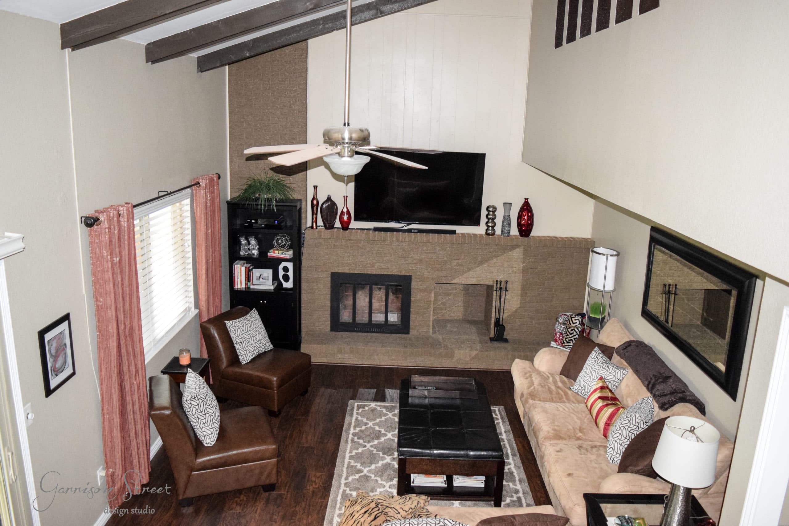 A cozy living room with a high ceiling and wooden beams. It features a stone fireplace with a TV above, surrounded by armchairs and a sofa with patterned cushions. A large mirror and shelves with decorative items enhance the space.