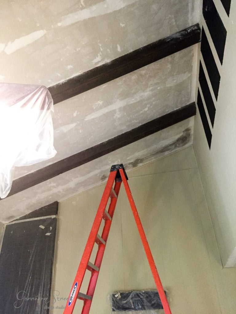 A room with a high ceiling featuring exposed wooden beams. There is a red ladder leaning against the wall. Some areas on the ceiling and walls are covered with plastic sheets, indicating a construction or renovation project.