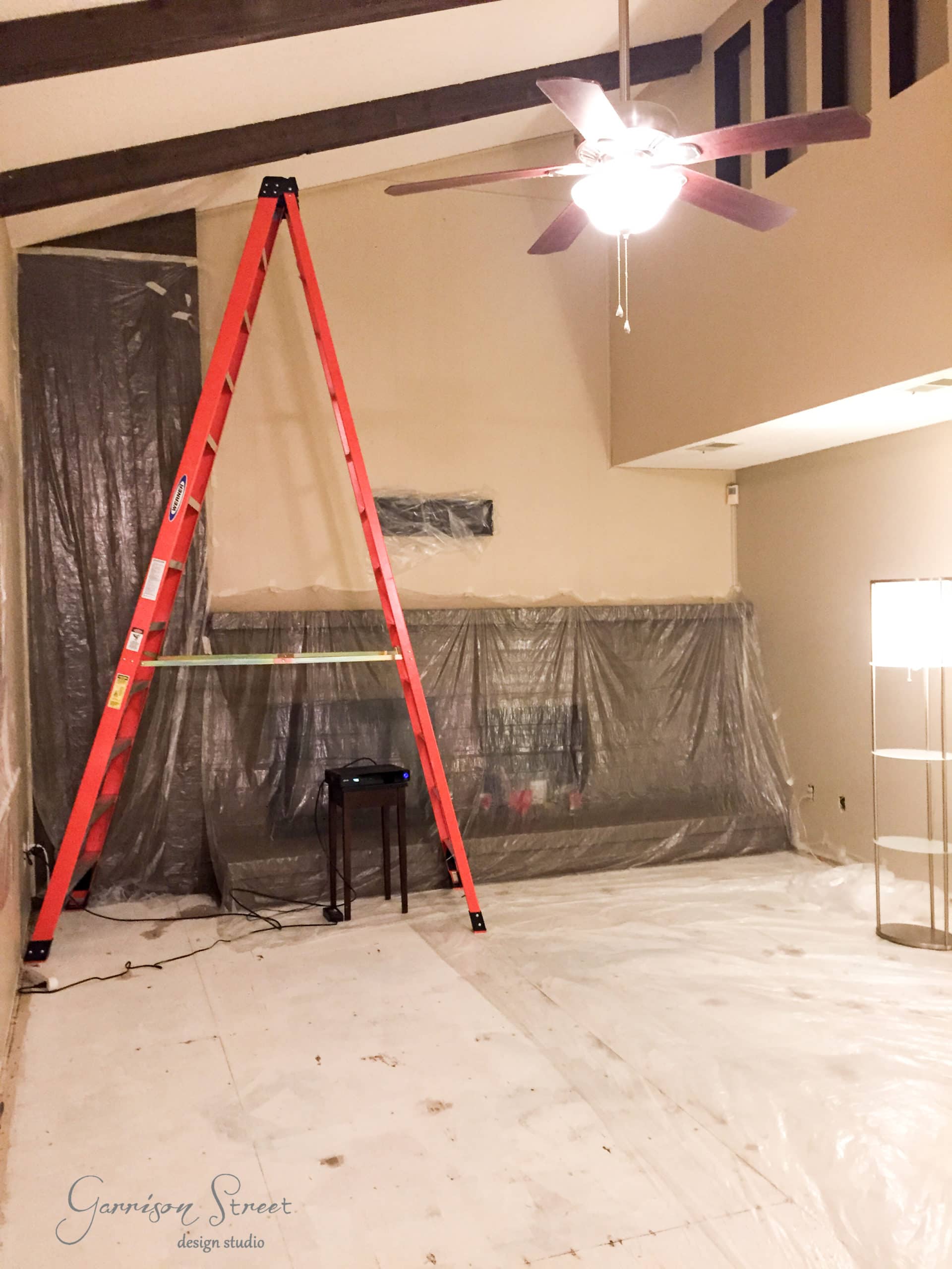 A room under renovation with a red ladder, covered furniture, and a plastic sheet on the floor. A ceiling fan with a light is on, and a standing lamp is in the corner. The walls and ceiling appear partially painted.