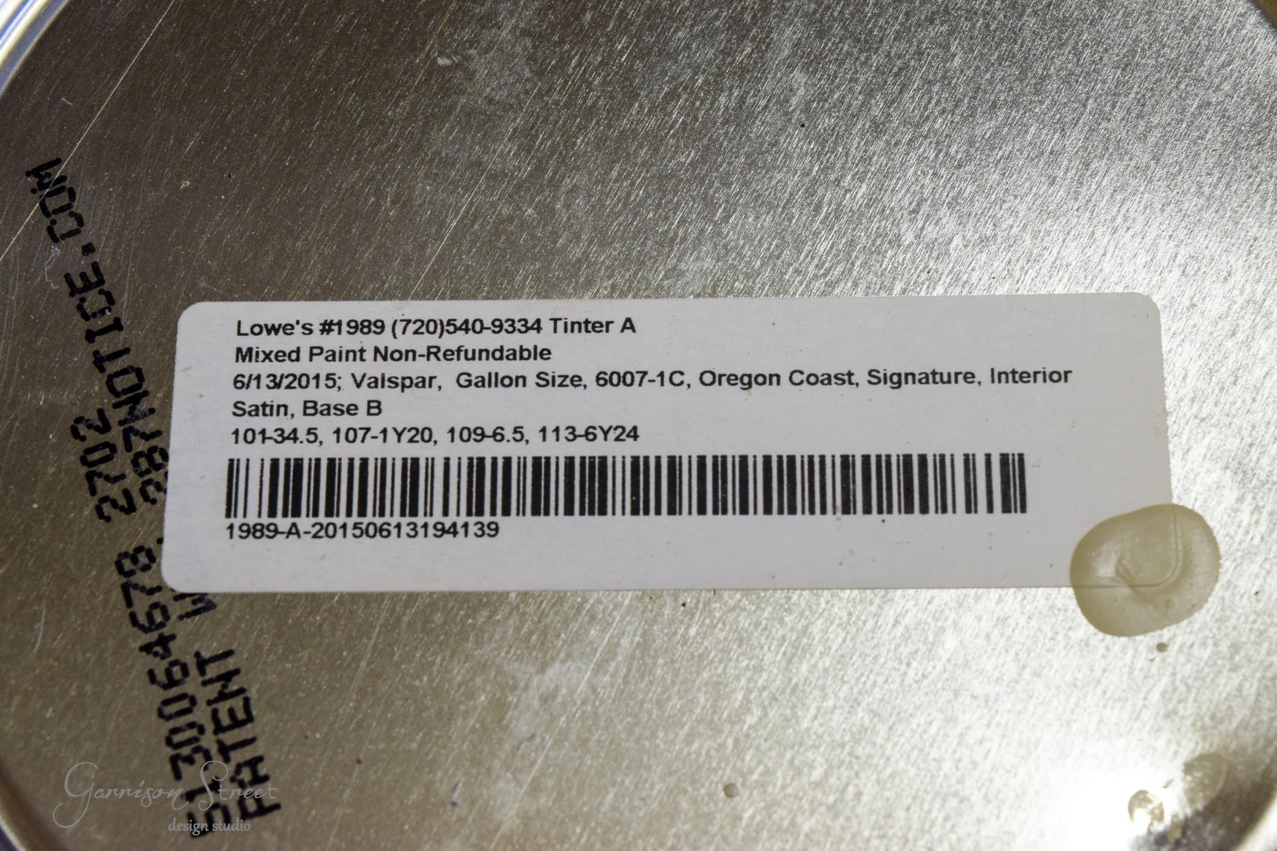Close-up of a paint can lid with a label showing details including Lowe's store number, date, Valspar paint, gallon size, color code (6007-1C), color name (Oregon Coast), finish (Satin), and base (Base B). Non-refundable notation included.