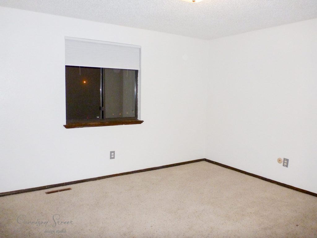 An empty room with white walls and a beige carpet. A single window with a closed blind is on one wall. The baseboards and window trim are dark wood, and there are two electrical outlets in the room.