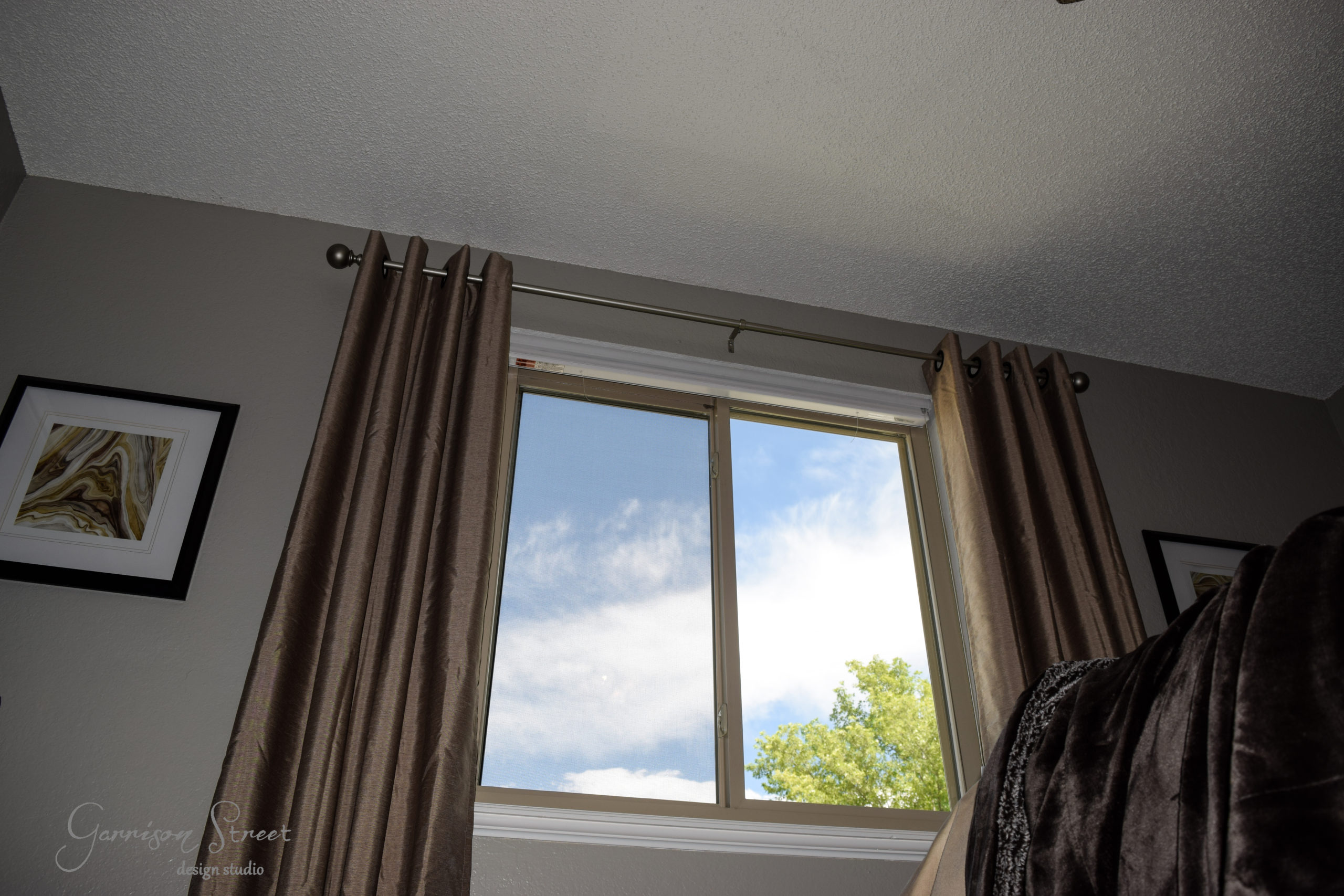 A window with open beige curtains revealing a view of a blue sky with a few clouds and a partial tree. The wall is gray with two framed pictures hanging beside the window. A part of a dark bedspread is visible in the foreground.