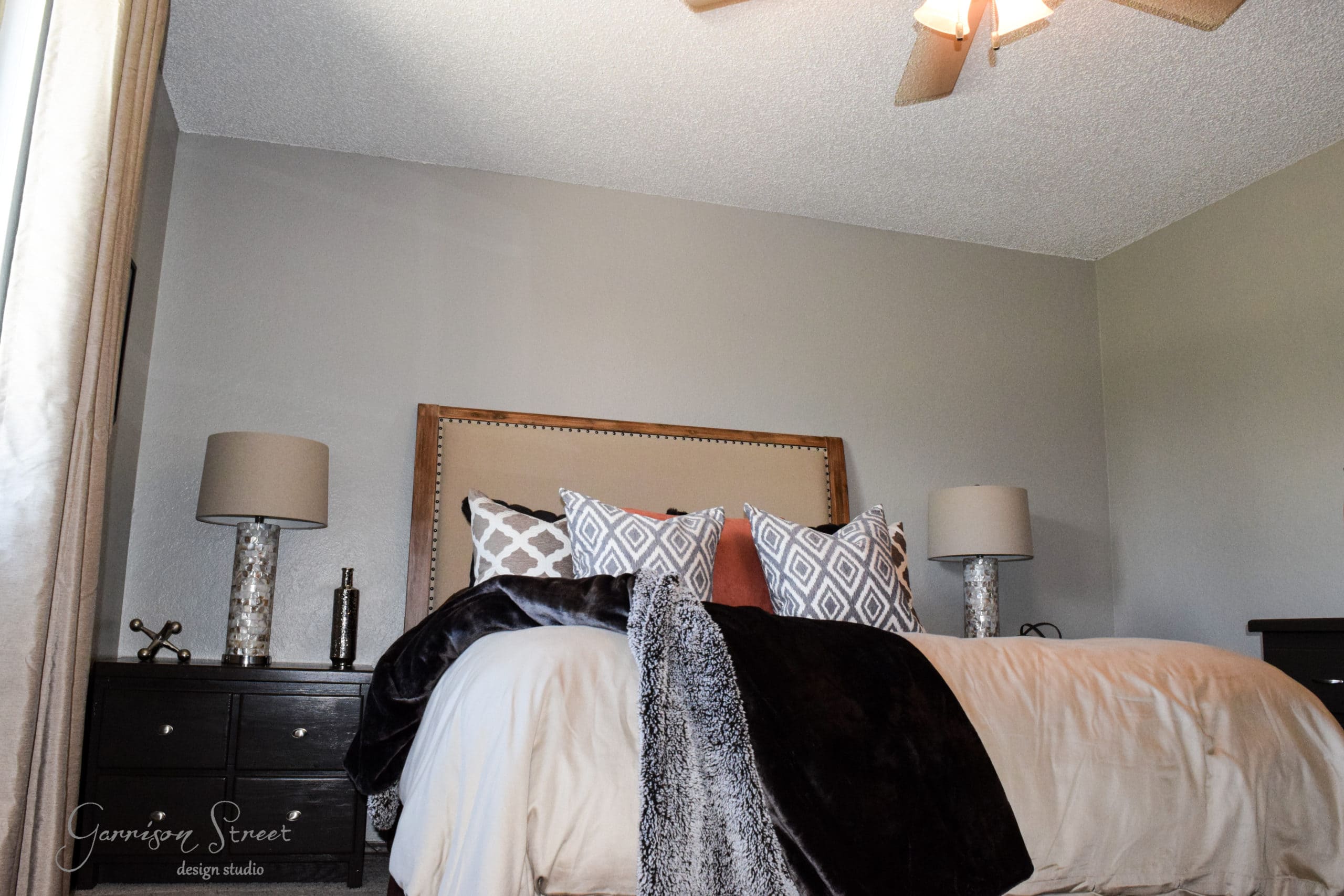 A bedroom with a large bed featuring patterned pillows and a dark throw. Two side tables with lamps flank the bed. The walls are light gray, and a ceiling fan is visible above. A window with curtains is partially seen on the left.