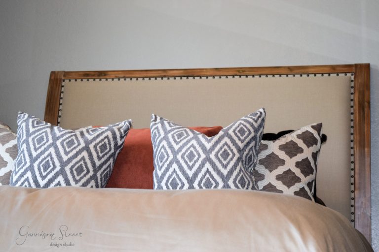 Bed with upholstered headboard, featuring three patterned throw pillows in black, white, and brown geometric designs, along with a brownish-orange accent pillow. The setting is minimal and modern.