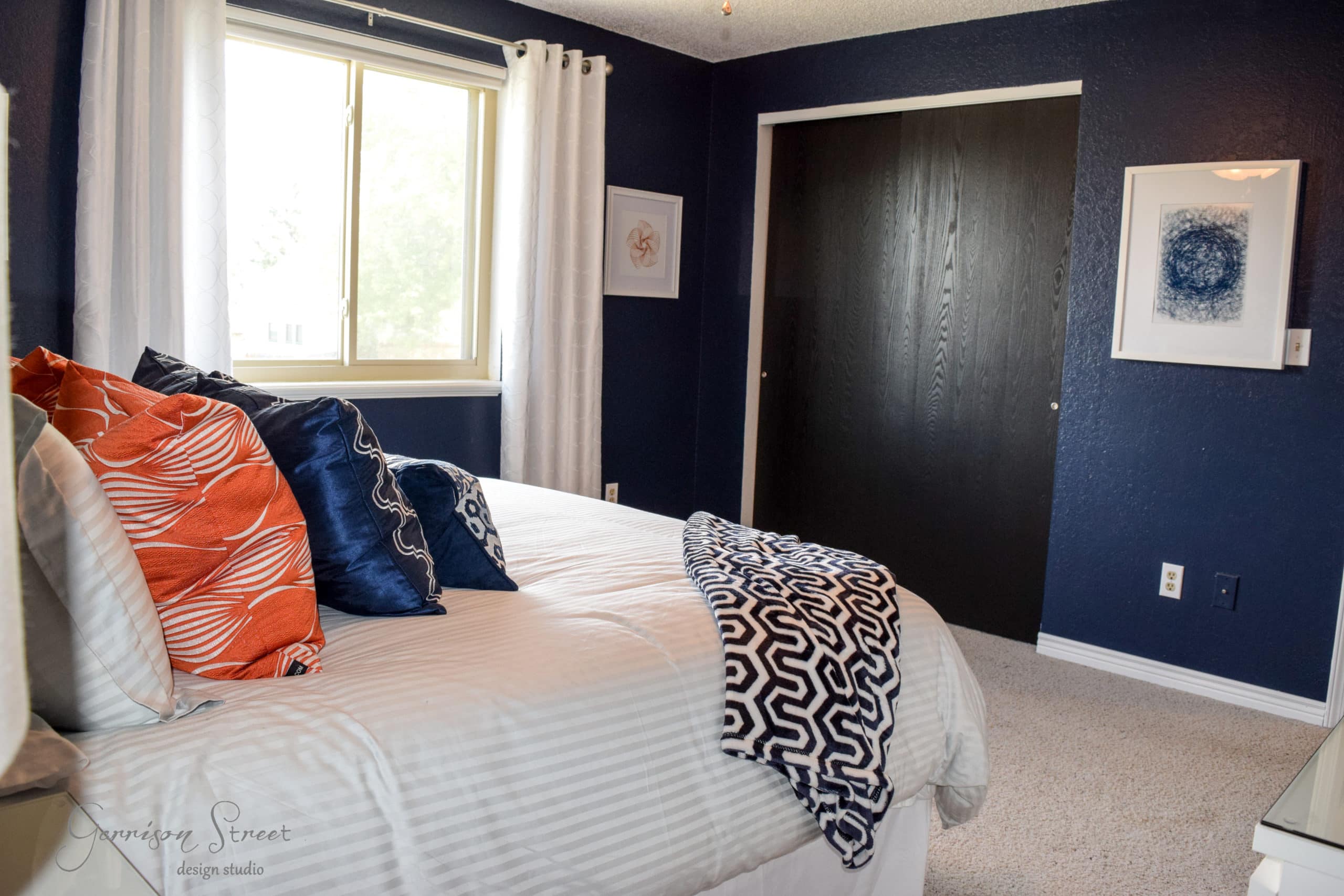 A bedroom with navy walls features a bed with white linens, orange and blue patterned pillows, and a navy patterned throw. A window with white curtains is on the left, and two framed artworks hang on the walls. A dark-colored closet door is opposite the bed.