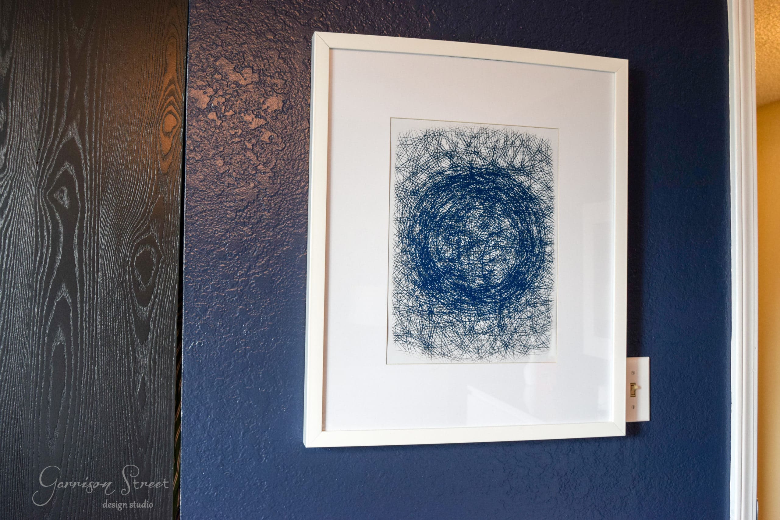 A framed abstract art piece with intricate blue spiral patterns hangs on a deep blue wall. A white doorframe is partially visible on the right side.