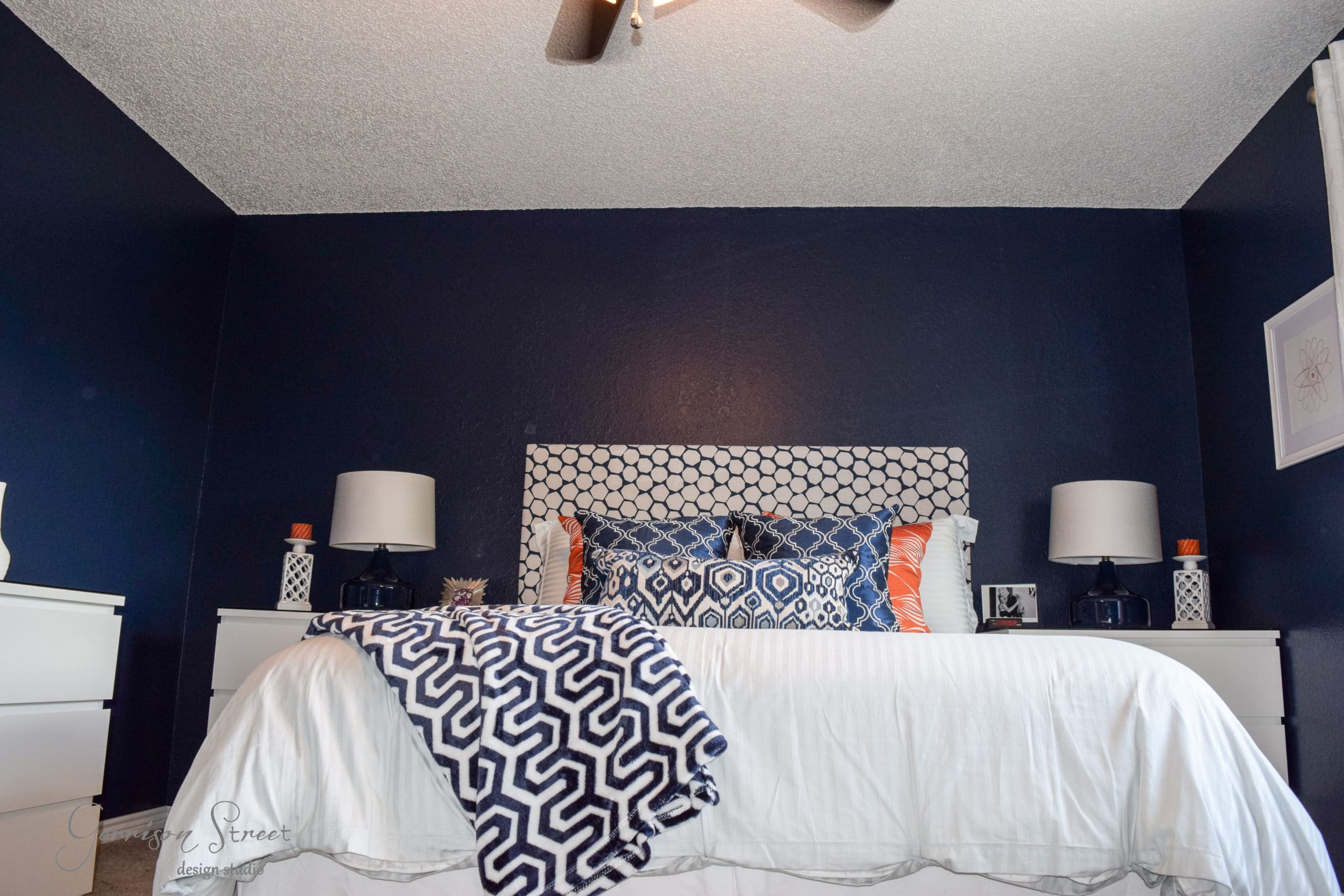 A bedroom with a navy blue accent wall features a bed with a geometric patterned headboard and matching bedding. Two white nightstands flank the bed, each topped with a lamp. Decorative pillows and a patterned throw blanket add contrast.