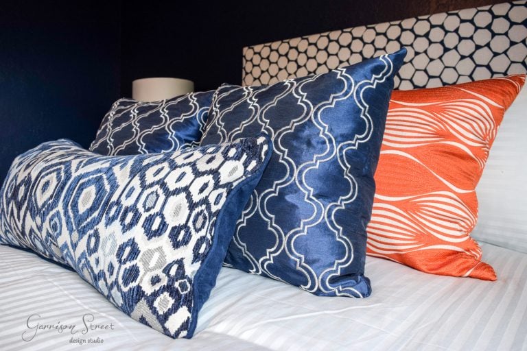 A bed with a decorative arrangement of three pillows: two with blue and white geometric patterns, and one with an orange and white wavy pattern. The background includes a headboard with a circular pattern.