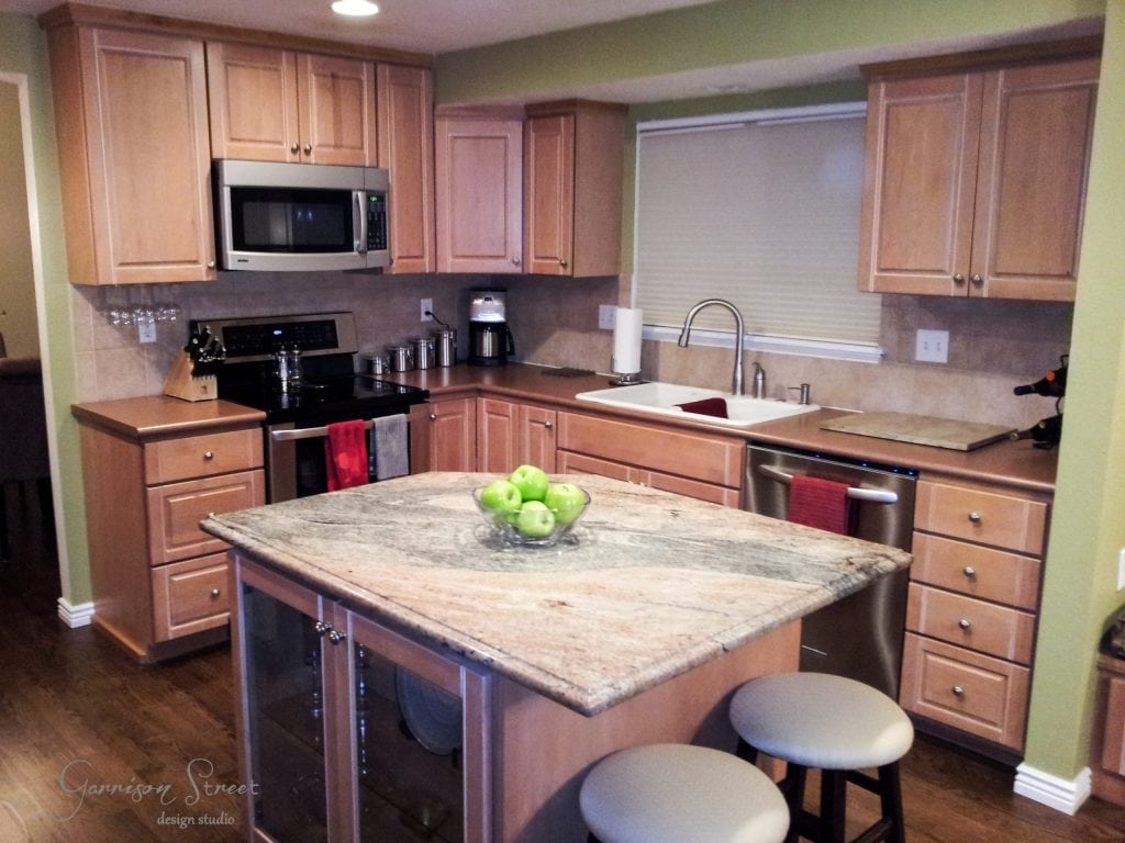 A modern kitchen with wooden cabinets and a granite island countertop exudes elegance after a dated 90s kitchen makeover. It features stainless steel appliances, including a microwave and stove. Green apples in a glass bowl sit on the island, with two stools nearby. Red towels hang by the sink.