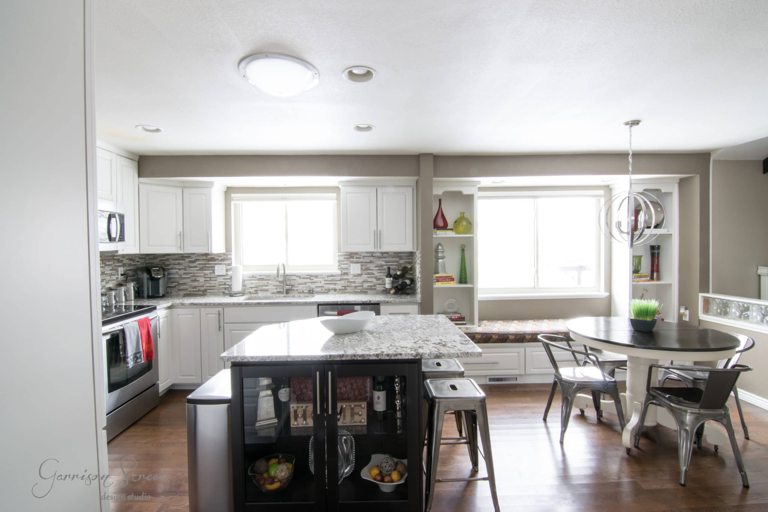 Bright and inviting, this modern kitchen showcases a stunning makeover from its dated 90s origins. Featuring sleek white cabinets, a central island with bar stools, and a cozy dining area with metal chairs around a round table, expansive windows flood the space with natural light over the polished wood floor.