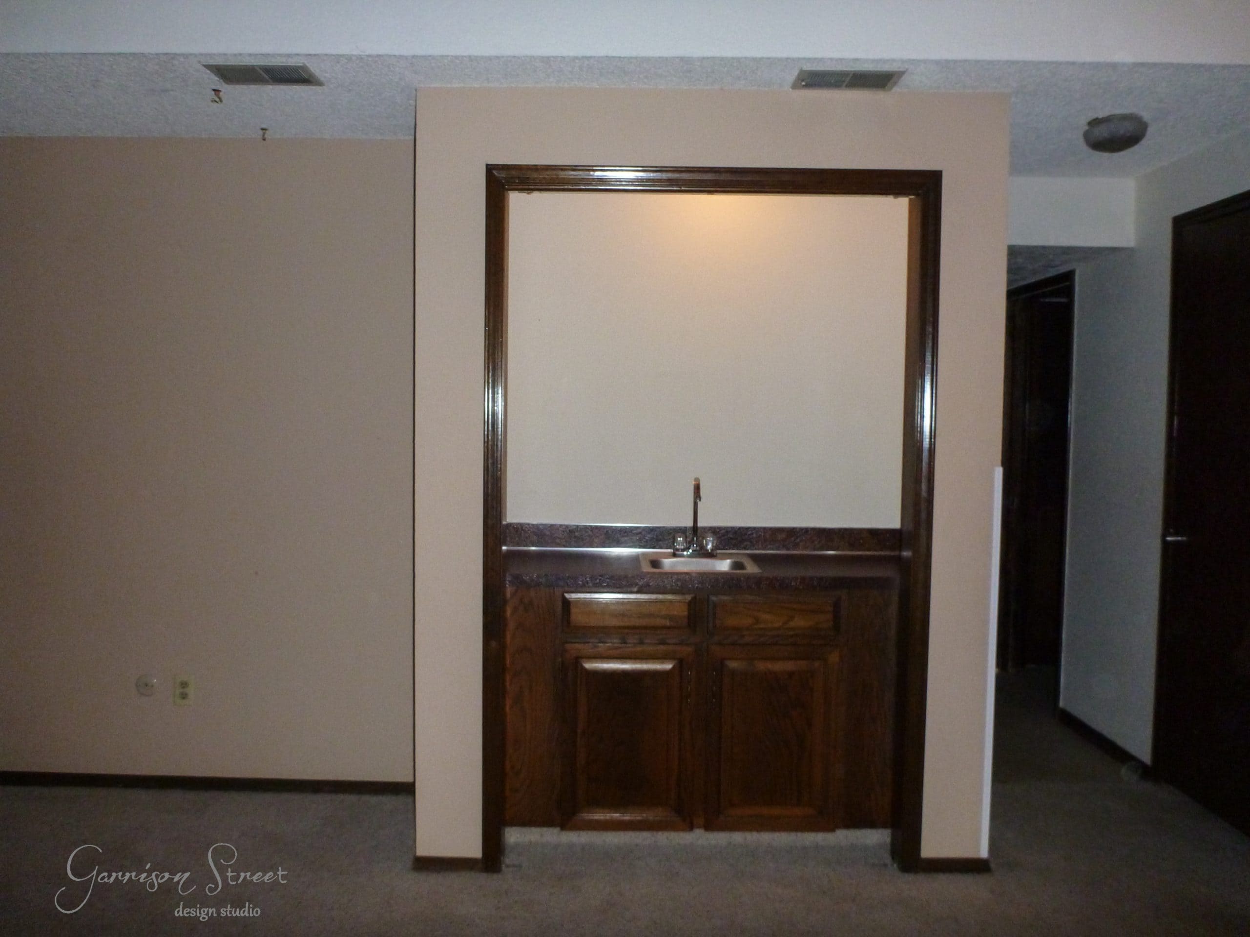 A small alcove with a built-in wooden cabinet and sink, surrounded by beige walls and a dark brown trim. The area is lit by a ceiling light, casting shadows on the wall and carpeted floor.