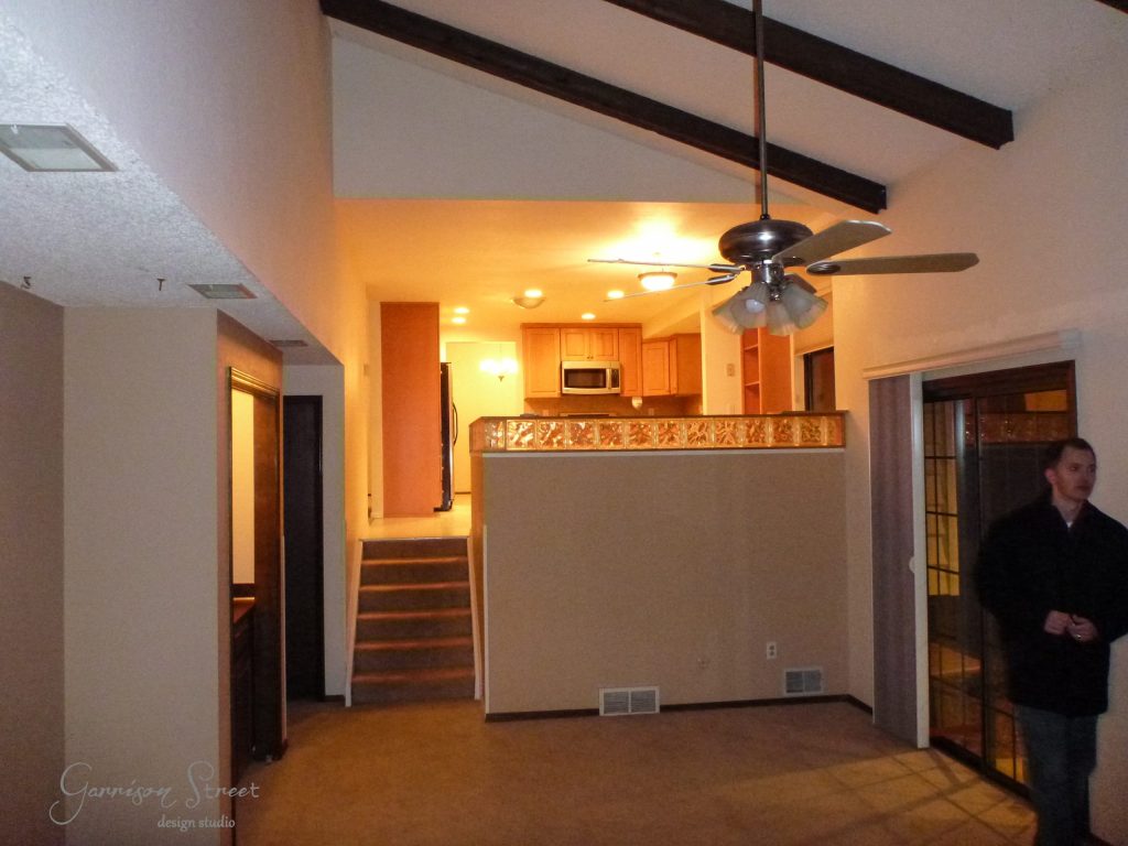 Interior of a home featuring a split-level layout. The lower area has beige carpet and walls, with a ceiling fan. Steps lead to a kitchen with wood cabinetry and recessed lighting. A sliding glass door is visible on the right, and a man stands near it.