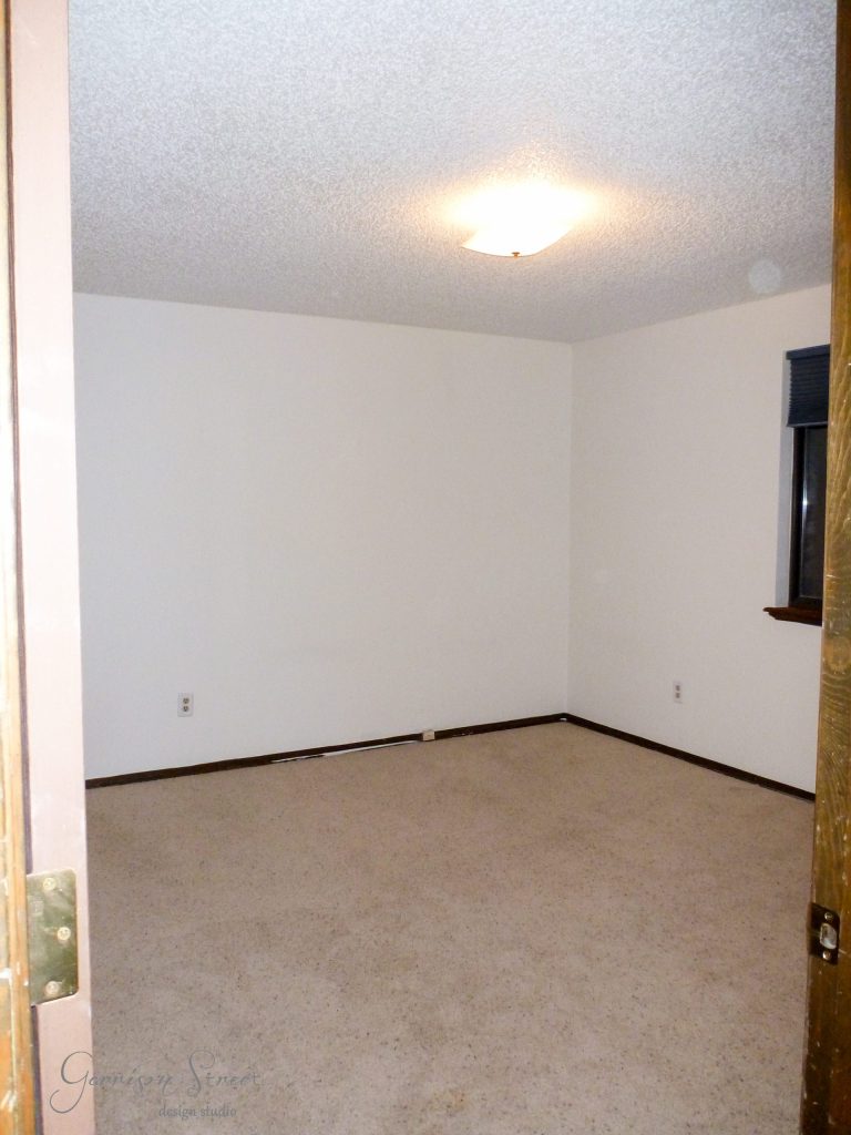 A small, empty room with beige carpeting and off-white walls. The ceiling has a textured finish and a ceiling light. There's a window with a closed dark shade and a wooden door frame visible on the left.