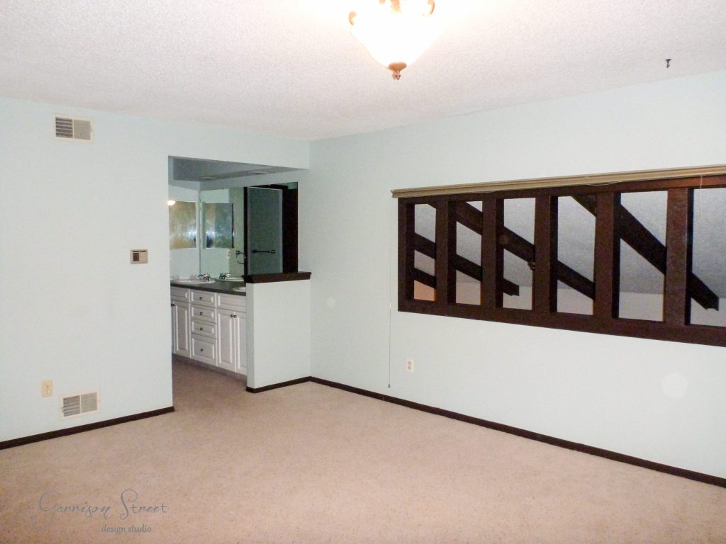 Empty room with light blue walls and beige carpet. A small nook with a sink and mirror is visible. The room has a ceiling light and a set of dark wooden louvered windows dividing it from another area.