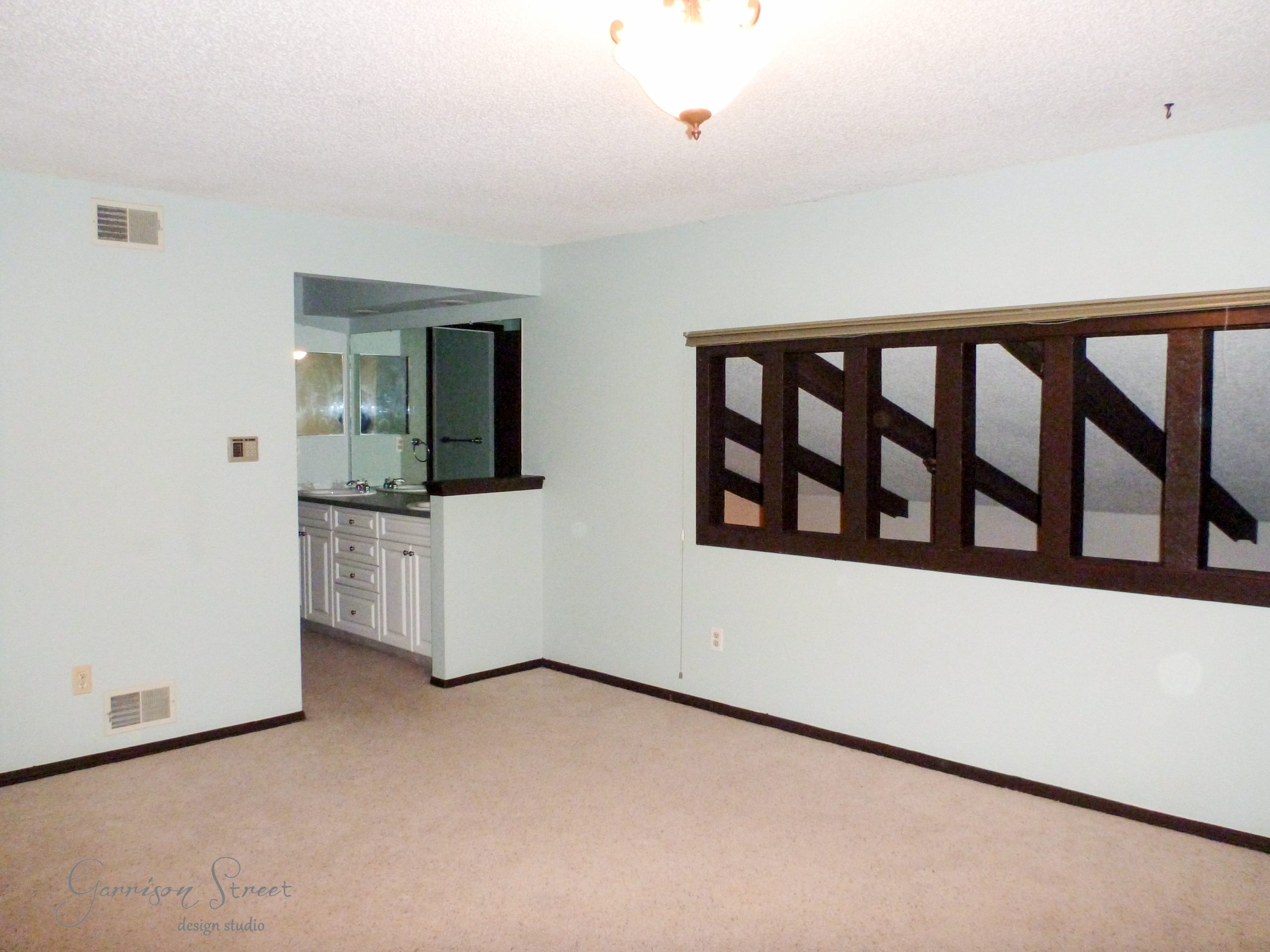 Empty room with light blue walls and beige carpet. A small nook with a sink and mirror is visible. The room has a ceiling light and a set of dark wooden louvered windows dividing it from another area.