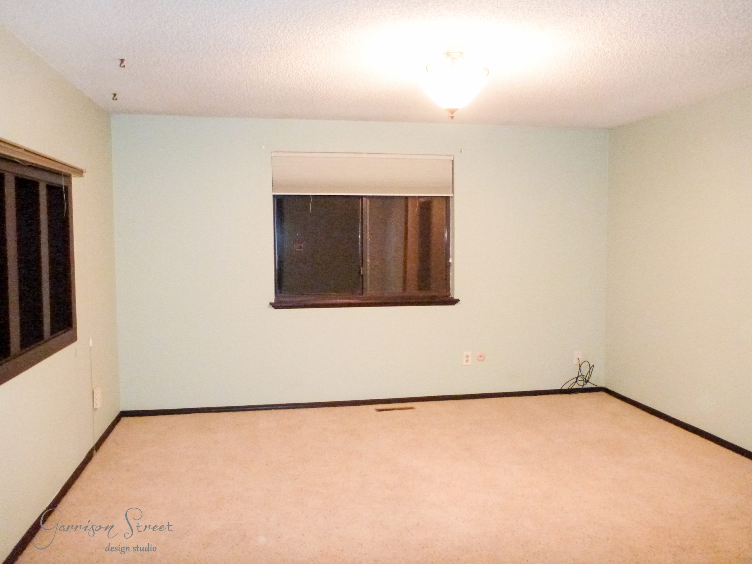 An empty room with light green walls, beige carpet, and a single ceiling light fixture. There are two windows, one with a blind partially down. The room has a minimalist design with no furniture.
