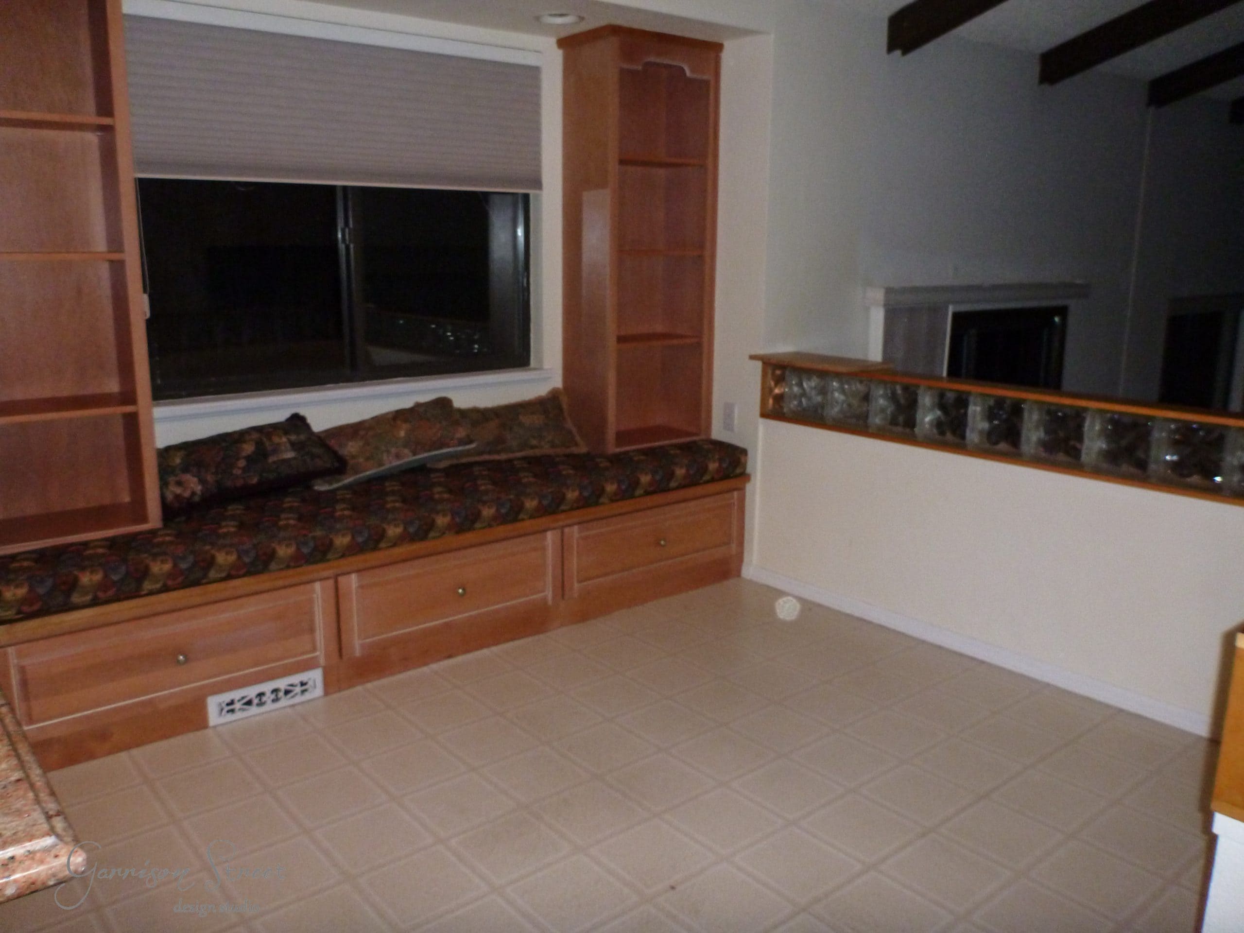 A living room with a built-in window seat featuring patterned cushions and wooden shelves. The room has tiled flooring, a glass block partition, and a large window with closed blinds. The area is dimly lit.