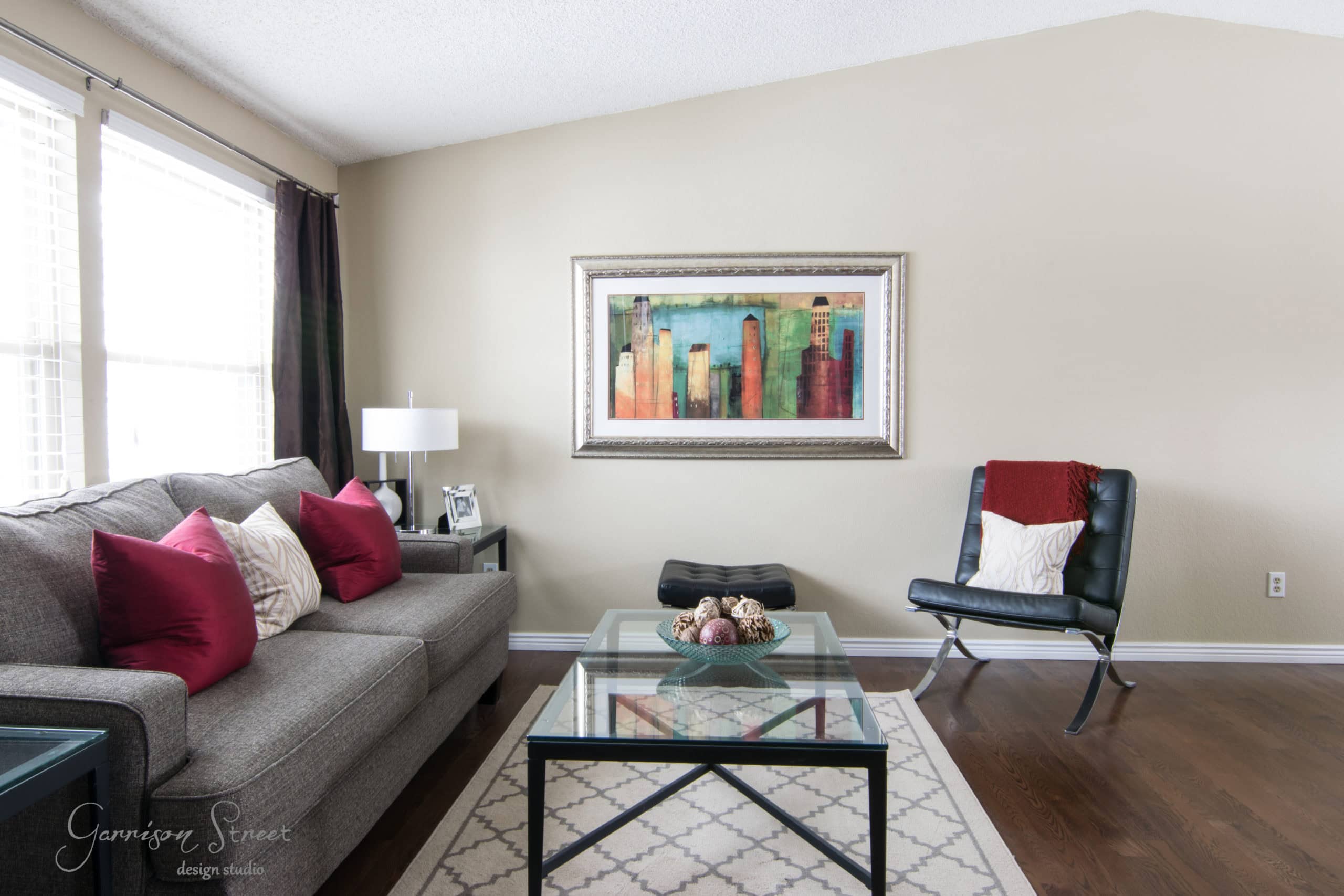 A modern living room with a gray sofa adorned with red and white cushions. A glass coffee table with decorative items sits on a patterned rug. A leather chair is on the right, and a colorful cityscape painting hangs on the beige wall.
