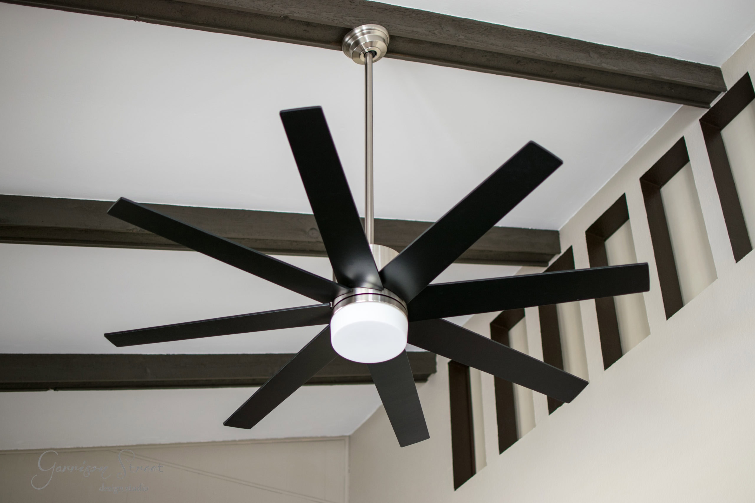 A modern ceiling fan with eight black blades and a silver base is mounted on a room ceiling. Dark wooden beams and sections of a wall with geometric cutouts are visible in the background.