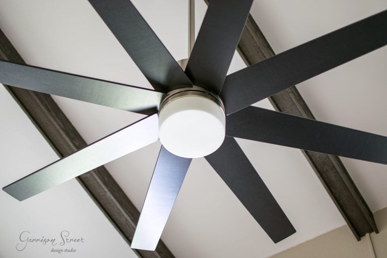 A large, modern ceiling fan with eight black blades is mounted on a ceiling with exposed wooden beams. The fan has a central white light fixture. The ceiling is painted in a light color, contrasting with the dark beams.