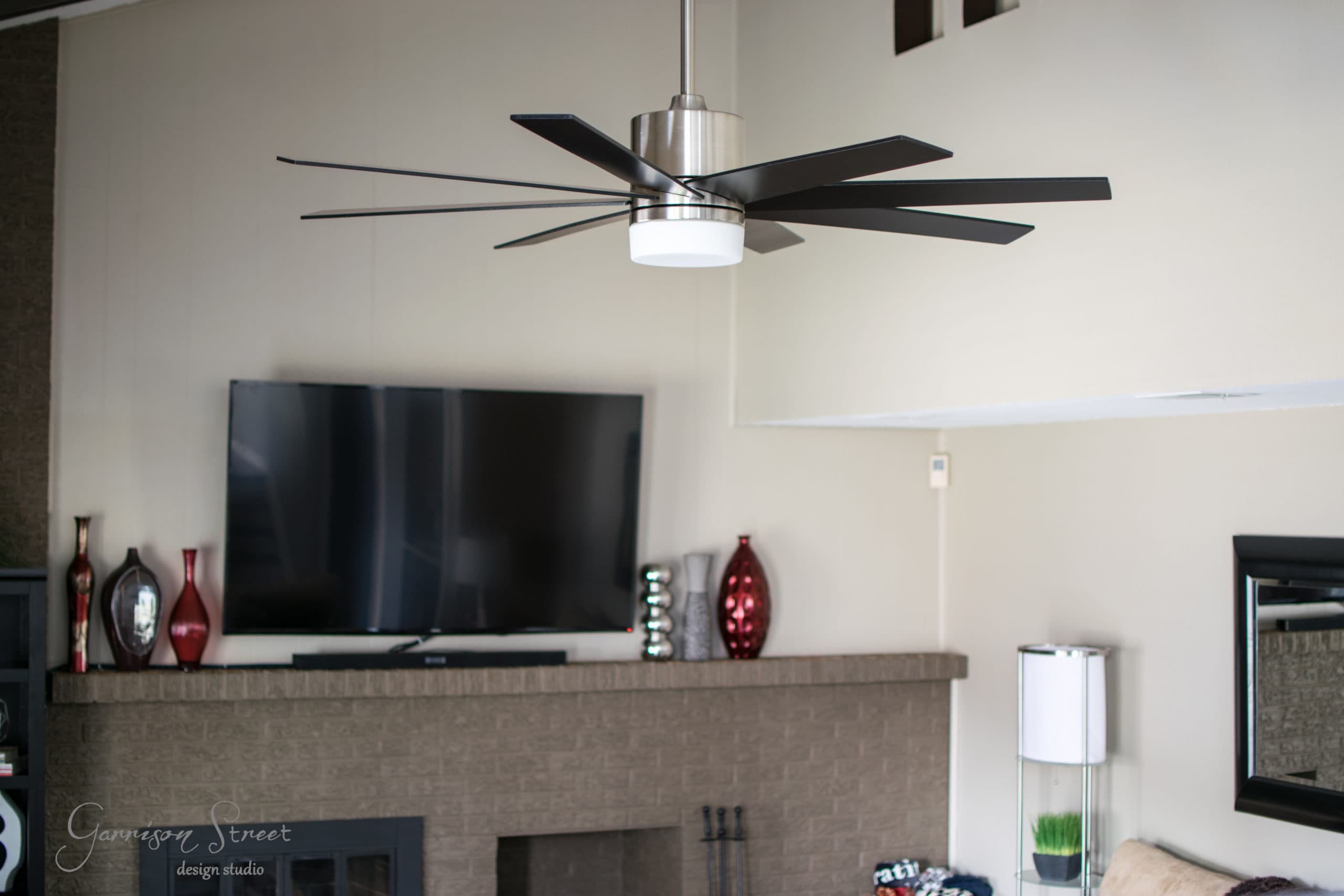 A modern living room with a large ceiling fan and dark blades. A flat-screen TV sits above a brick fireplace, flanked by red and silver decorative vases. Light-colored walls and a tall, cylindrical lamp are also visible.
