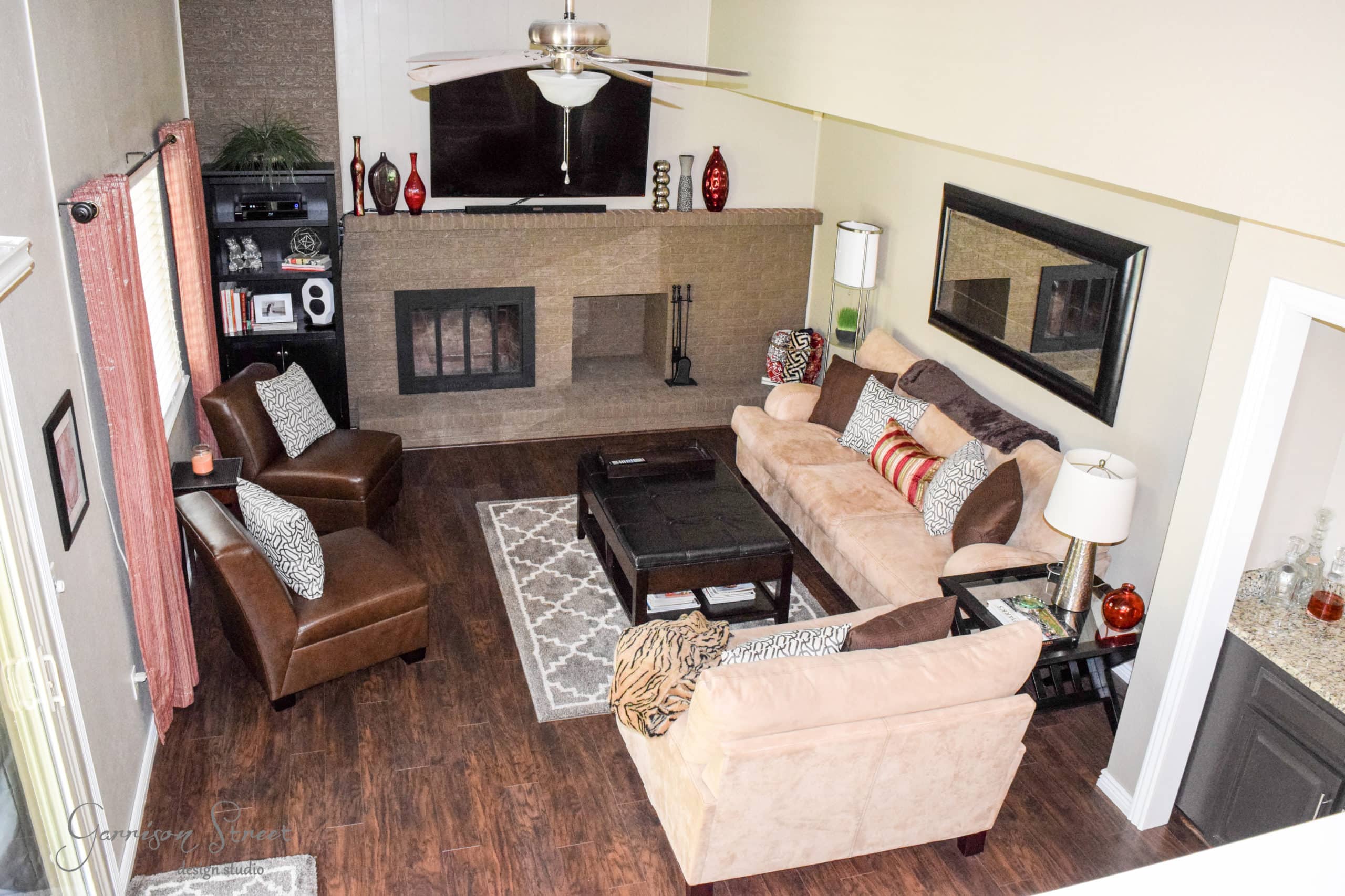 A cozy living room with dark wooden flooring, a large beige sofa, and two brown armchairs. A flat-screen TV is mounted above a fireplace. The room has decorative vases, a patterned rug, a mirror on the wall, and natural light coming from a window.