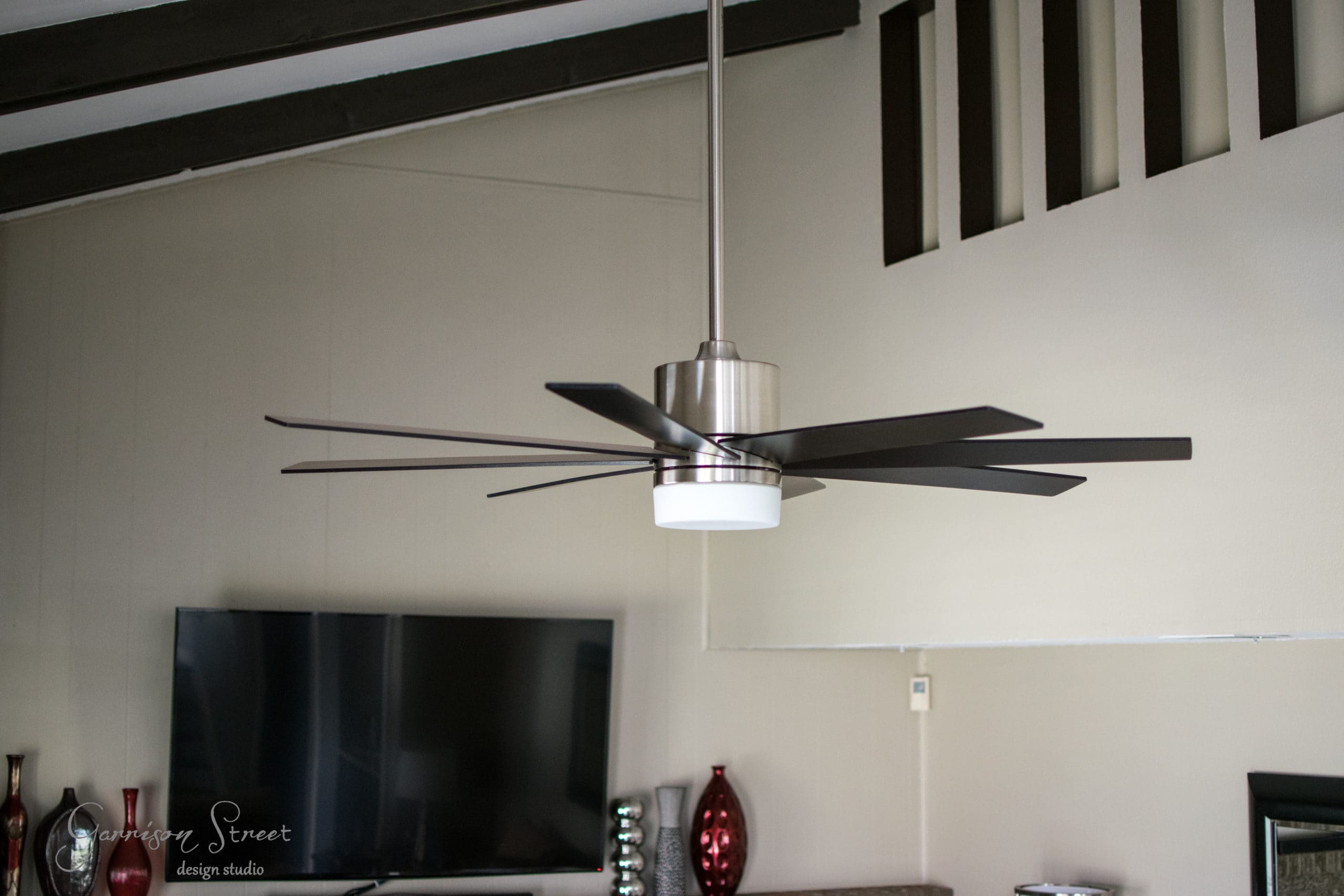 Modern ceiling fan with silver base and dark blades, centrally suspended in a living room with a TV mounted on the wall and decorative vases on a shelf. The room has a minimalist aesthetic with light-colored walls and a dark beam ceiling.
