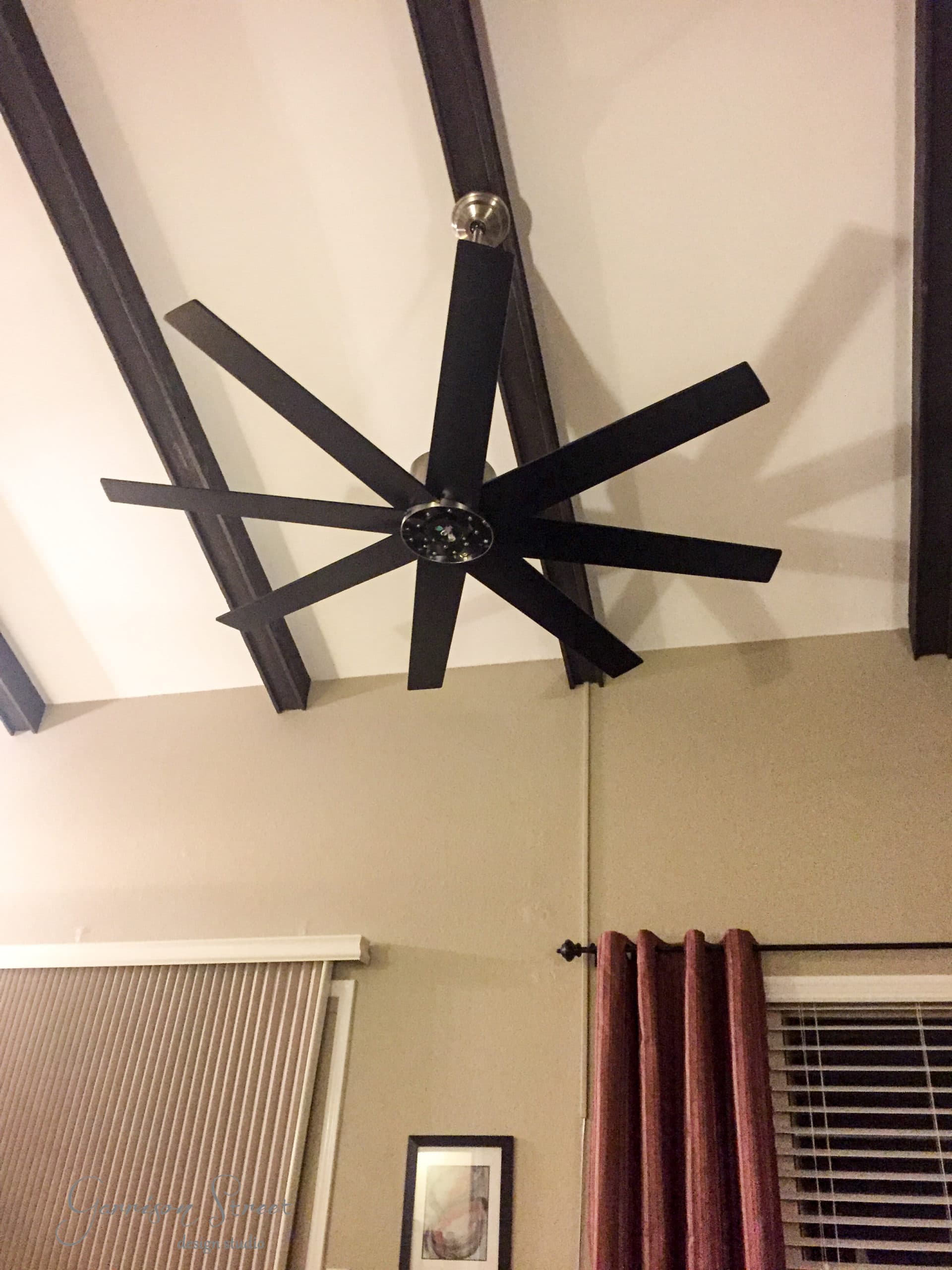 A large black ceiling fan with eight blades is mounted on a high, vaulted ceiling. Beams run parallel to the fan. Below are beige walls with a framed picture and a window covered with blinds and red curtains.