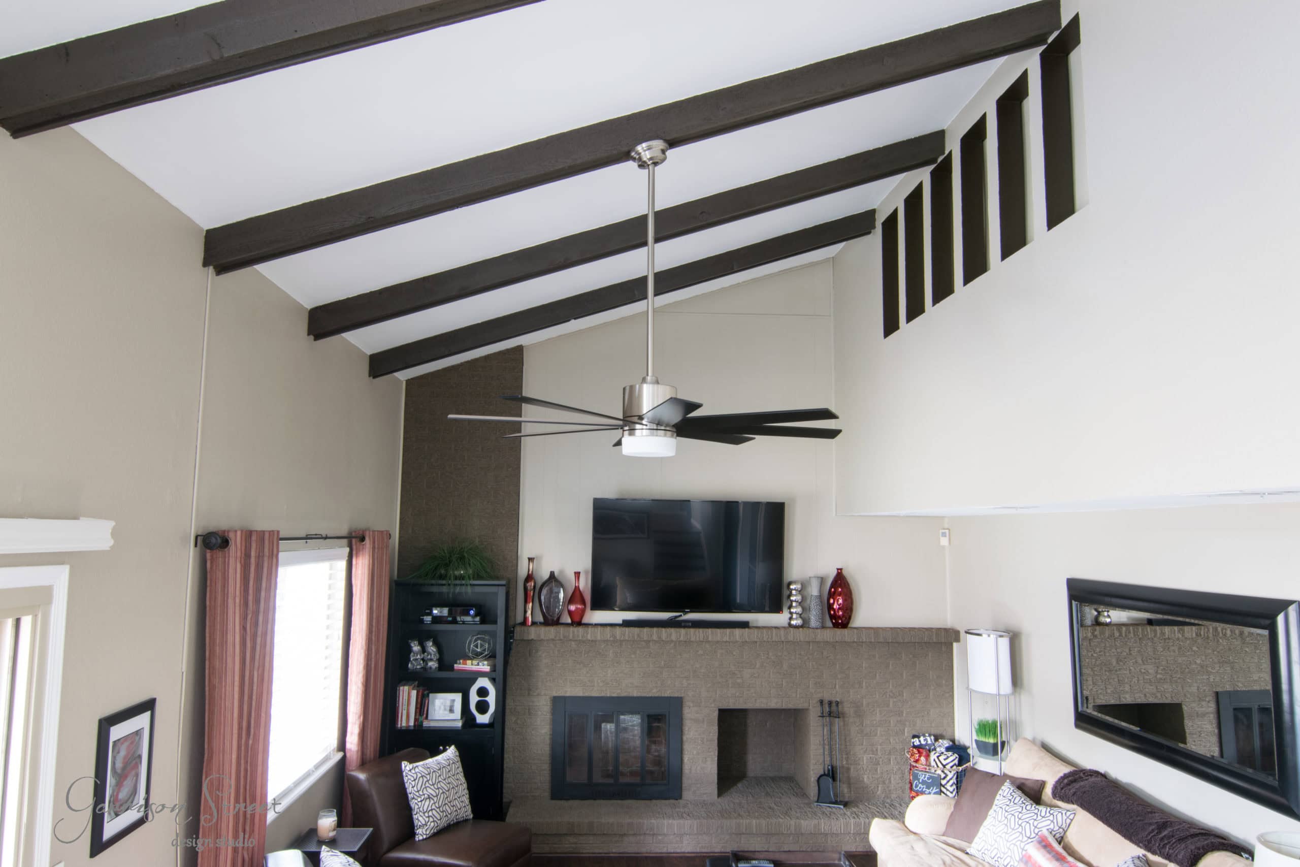 A modern living room with high ceilings and exposed wooden beams. Features a ceiling fan, a TV mounted above a brick fireplace, and a brown leather chair. Shelves with decorative items flank the fireplace, and there's a large mirror on the right wall.
