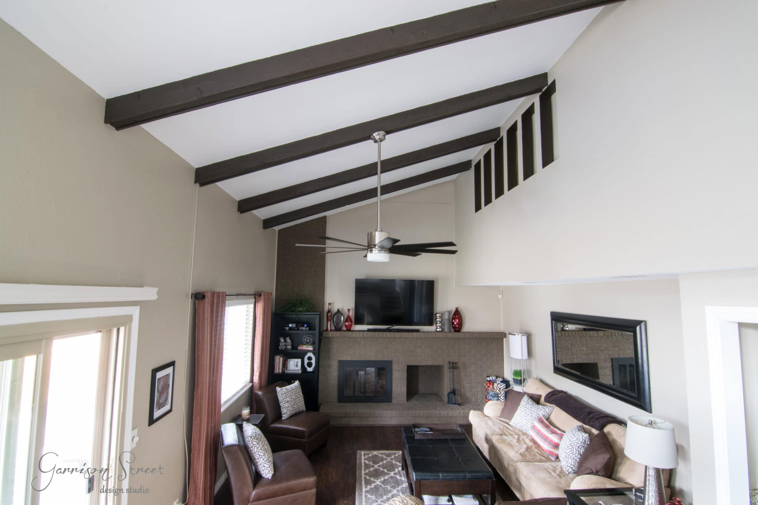 Spacious living room with high ceiling and exposed wooden beams. Features a ceiling fan, large TV above a fireplace, and comfortable seating around a coffee table. Neutral color palette with natural light from a window on the left.