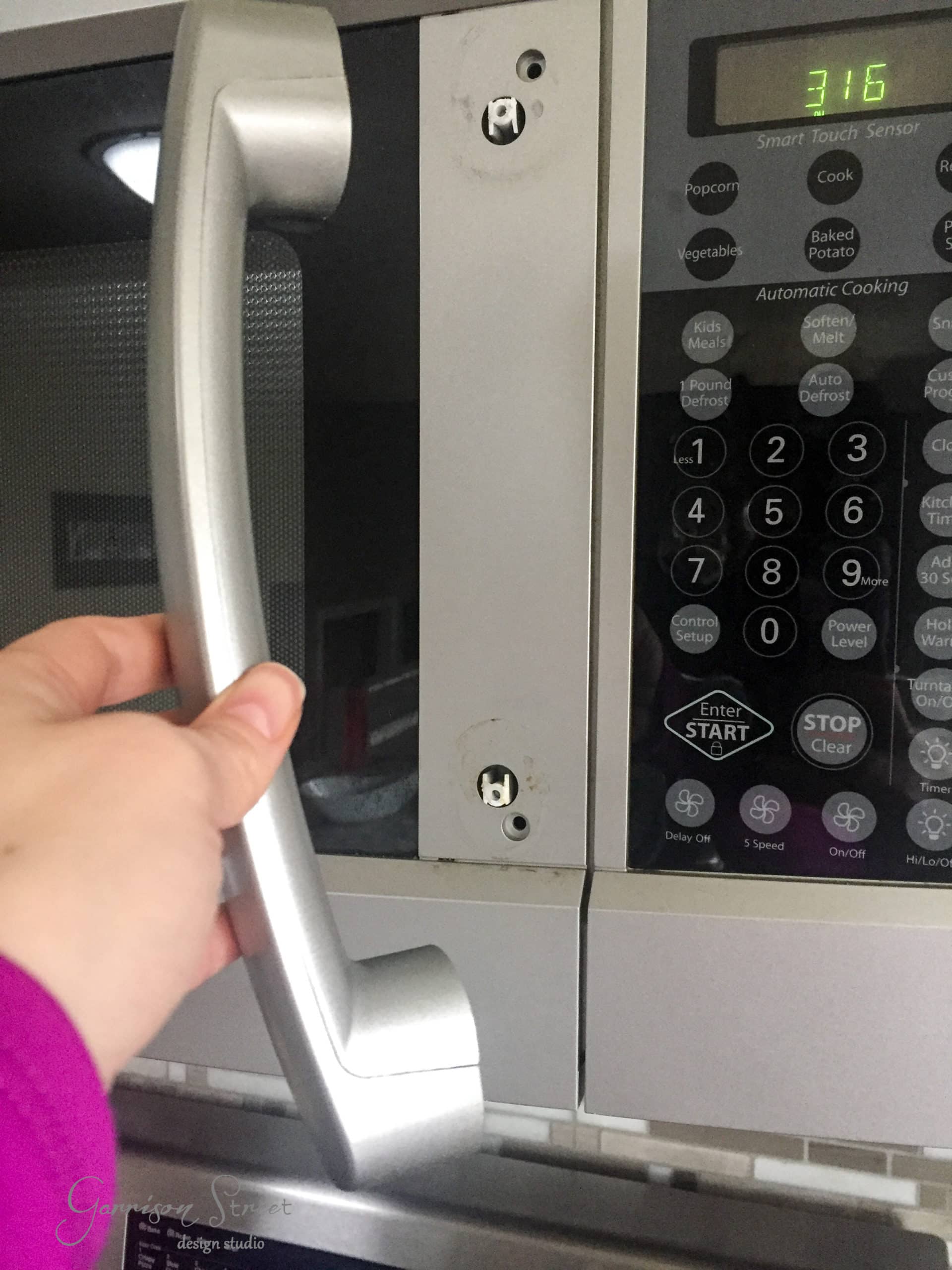 A person wearing a purple sleeve holds a detached microwave door handle near the microwave. The control panel displays "3:16." The handle is missing screws, and the door shows part of a Smart Touch Sensor panel with various buttons.