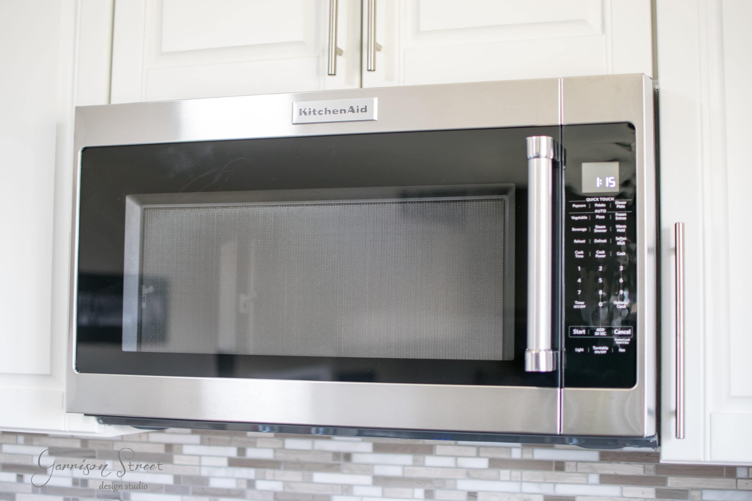 A stainless steel KitchenAid microwave with a digital display and control panel, set above a tile backsplash. The microwave is installed under white kitchen cabinets.