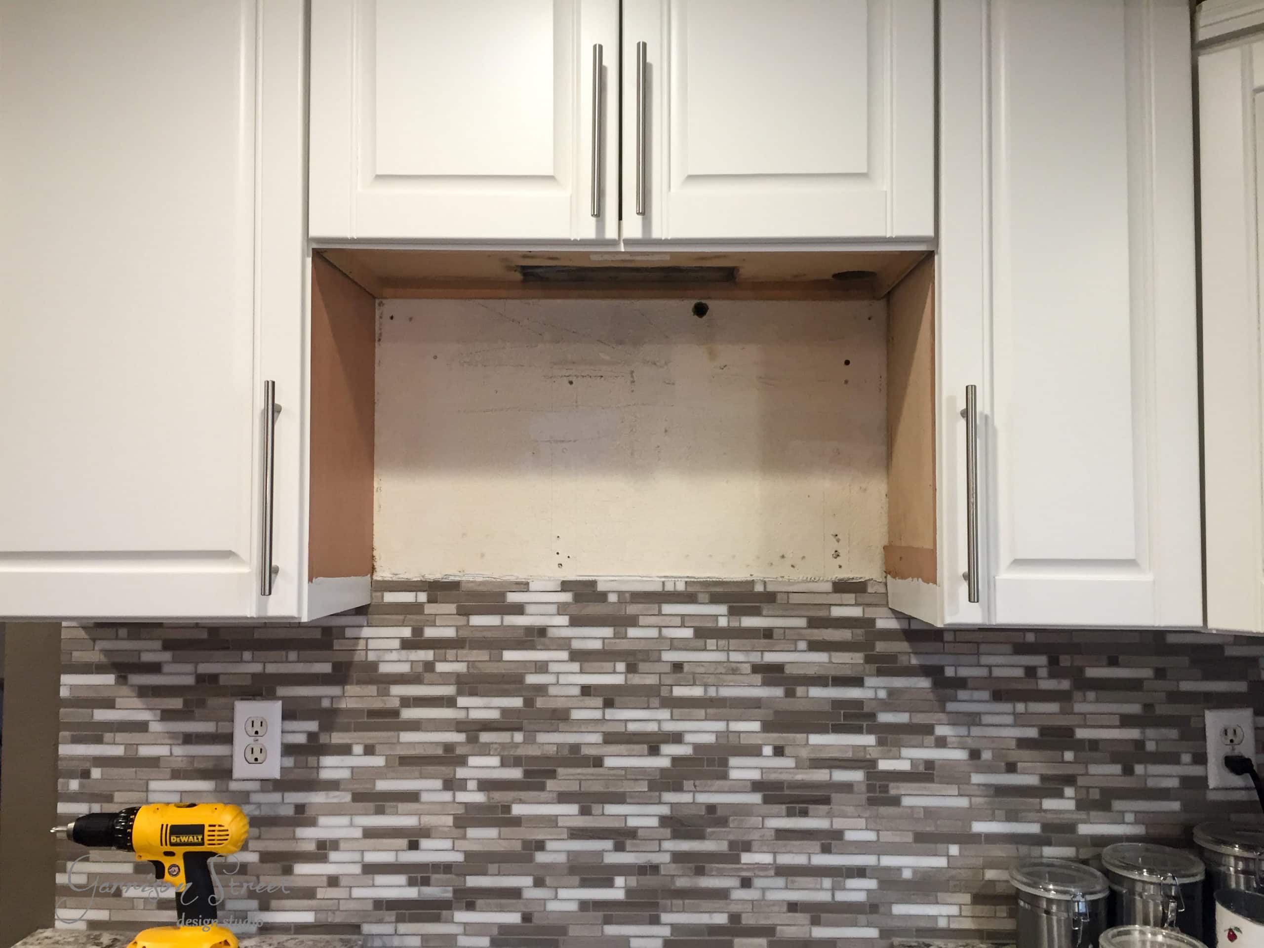A kitchen with white cabinets and a gray tile backsplash. The space above the countertop is empty, likely prepared for a range hood installation. A drill and some tools are placed on the counter.
