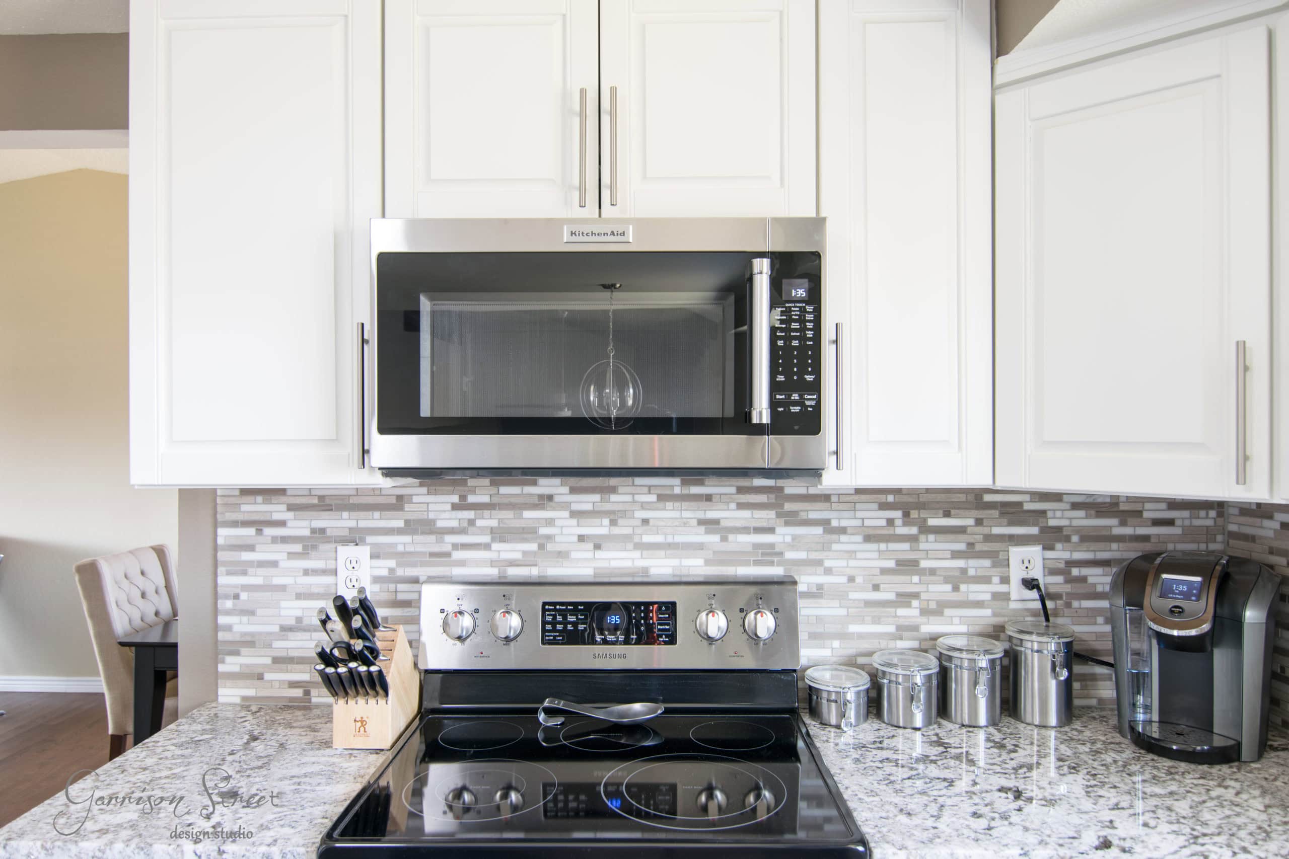 Modern kitchen with a stainless steel microwave above an electric stove, surrounded by white cabinets. Granite countertops hold a knife block, glass canisters, and a coffee maker. The backsplash features gray and white mosaic tiles.