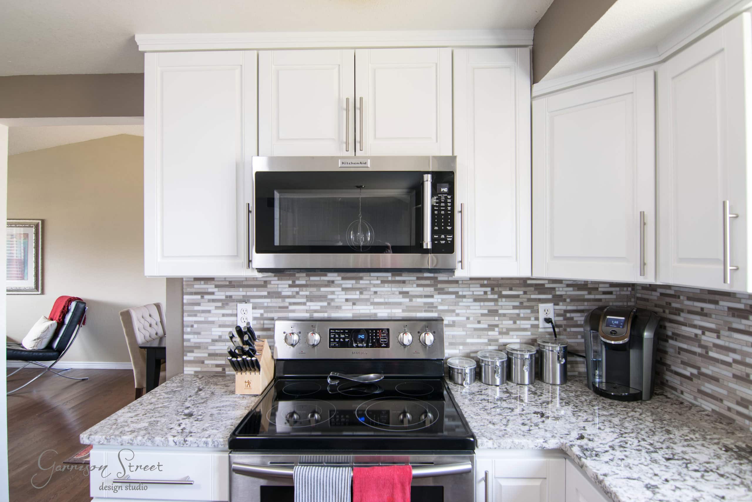 Modern kitchen with white cabinets, stainless steel appliances, and a granite countertop. A microwave is installed above the stove, and a coffee maker sits on the counter. Knives and canisters are arranged neatly nearby.
