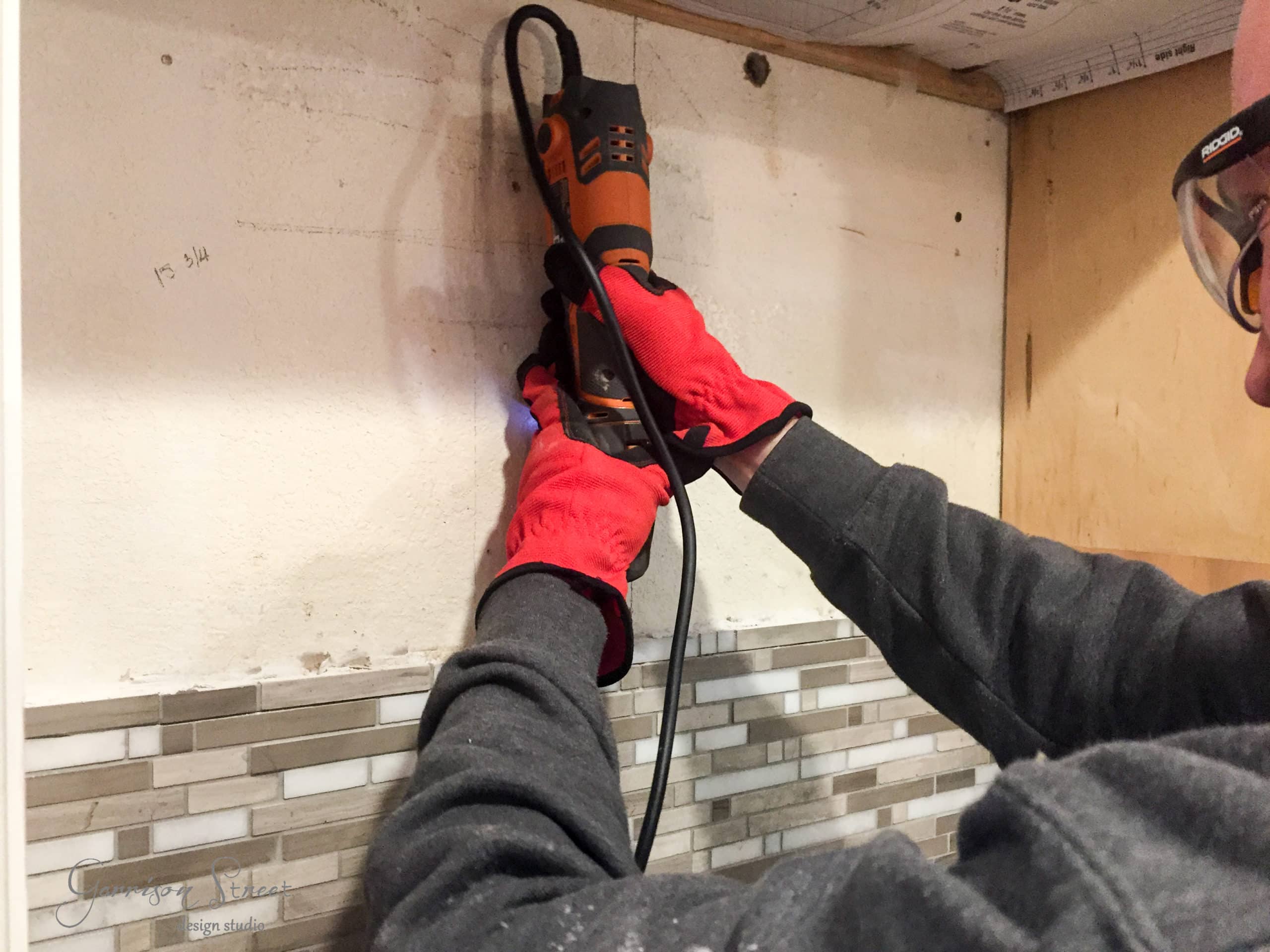 A person wearing red gloves and safety glasses uses a power tool to cut or sand a wall above a tiled backsplash in a kitchen.