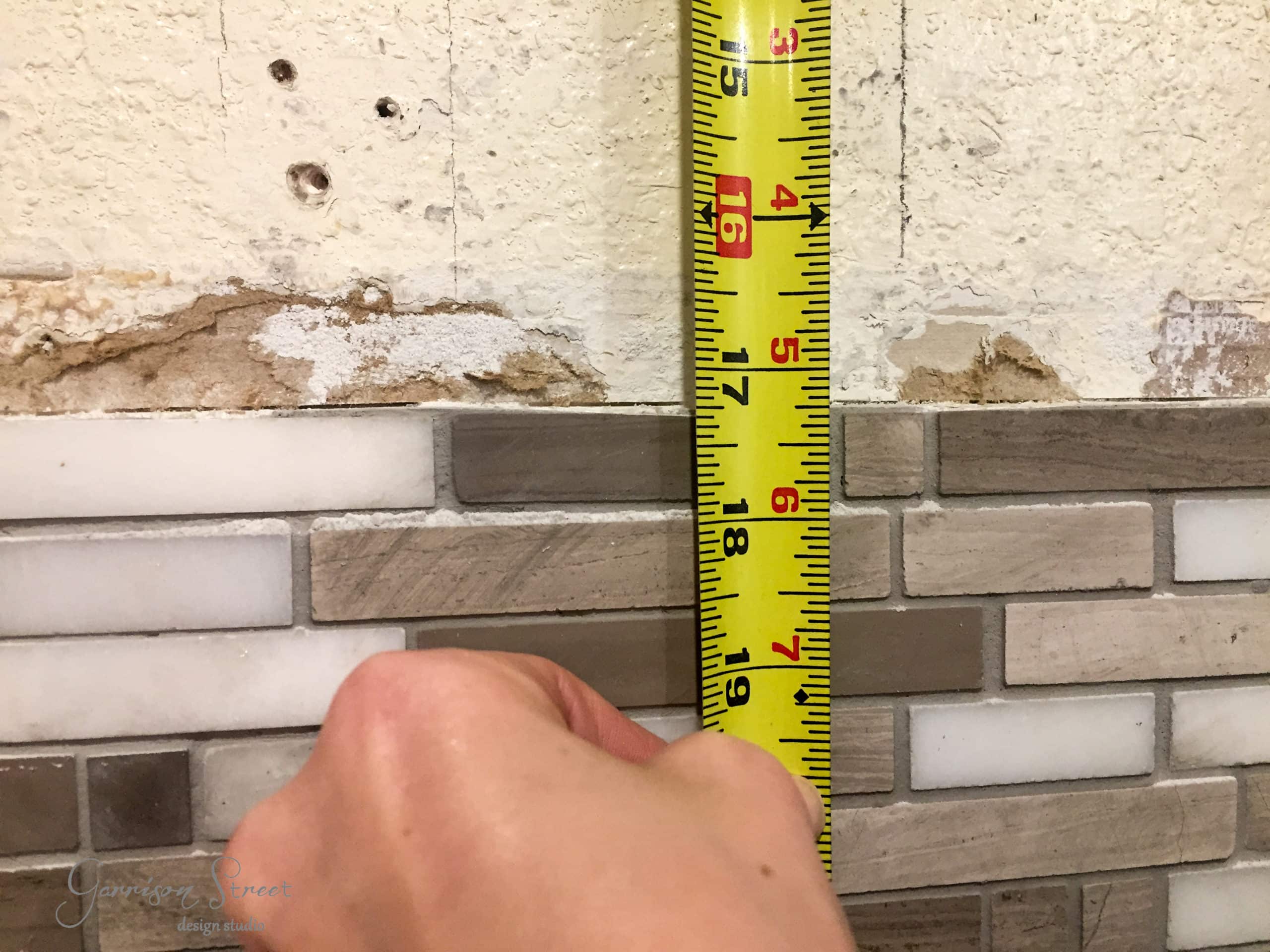 A hand holds a yellow measuring tape against a wall with beige and brown mosaic tiles. The wall above the tiles is unfinished, showing some wear and missing patches.
