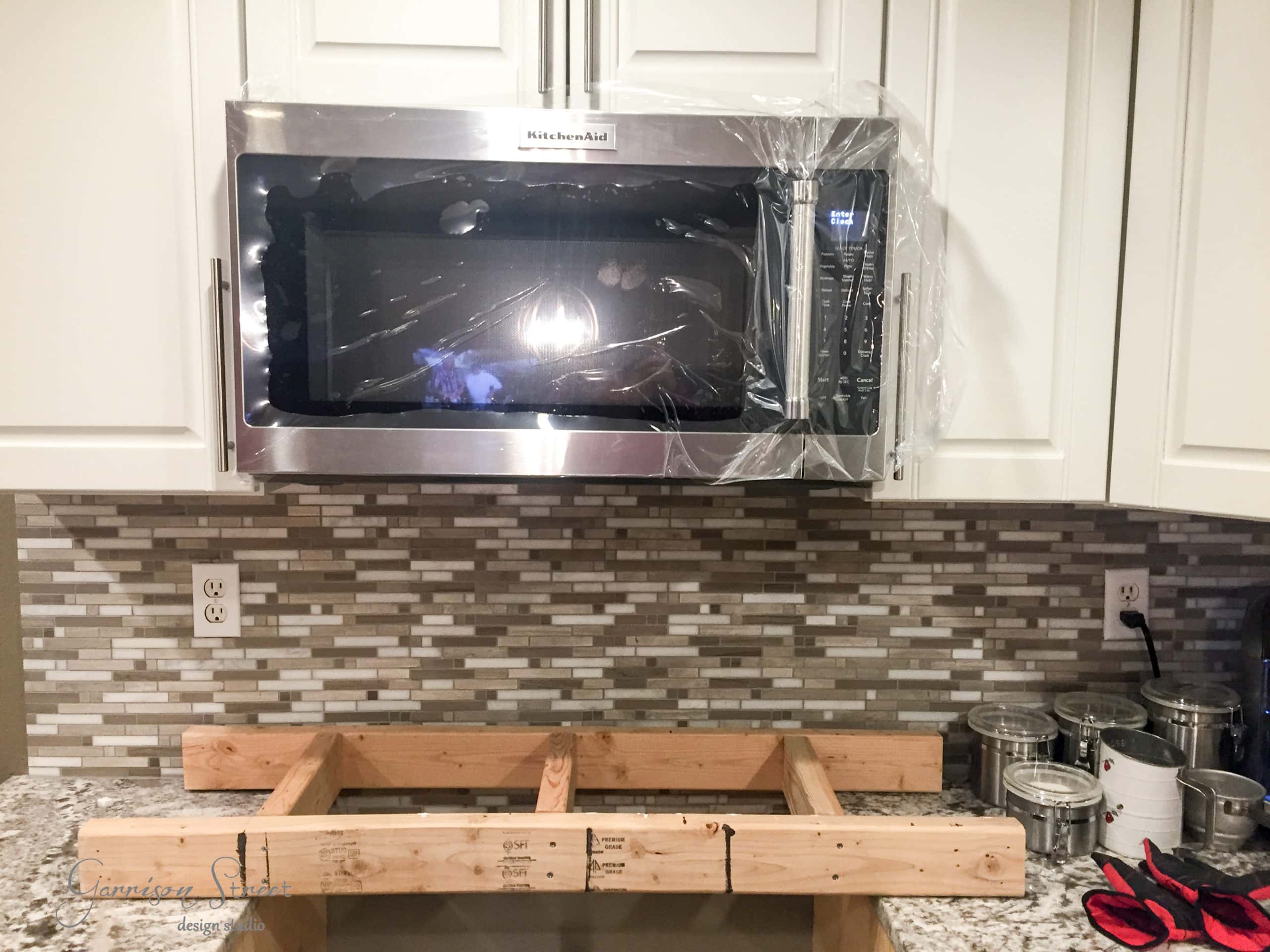 A new microwave, partially wrapped in plastic, is installed above a kitchen counter with a tiled backsplash. Below it, wooden planks are set up, possibly for support. A pair of red and black gloves are on the counter.