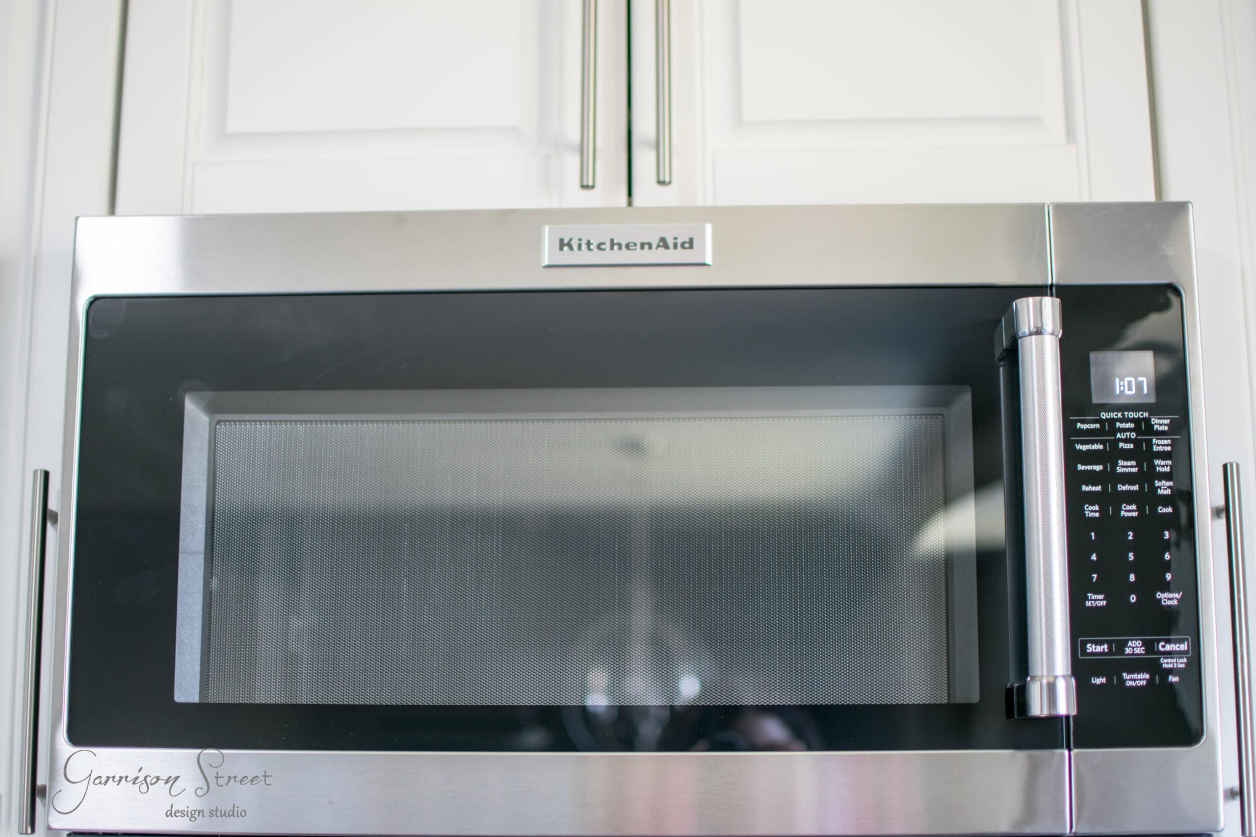 Close-up of a stainless steel KitchenAid microwave installed in a kitchen cabinet. The digital display shows the time as 1:07. The microwave has a handle on the right side and several buttons for different functions below the display.