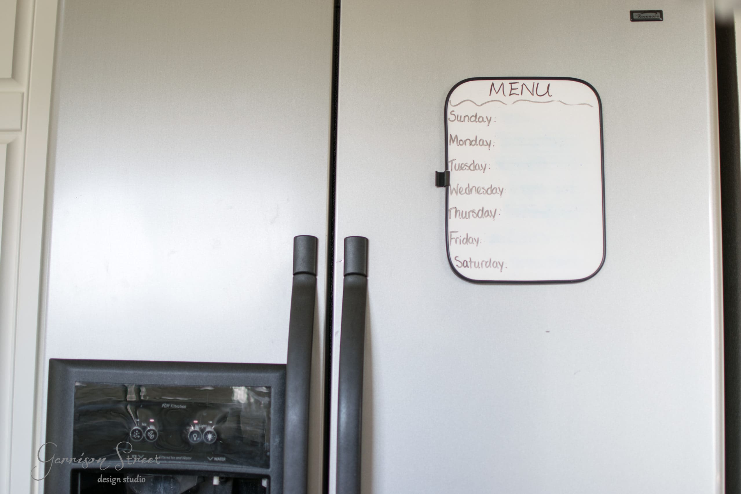 A stainless steel refrigerator with black handles. A white dry erase board is attached to the door, displaying the days of the week from Sunday to Saturday, titled "Menu". The freezer has an ice and water dispenser.