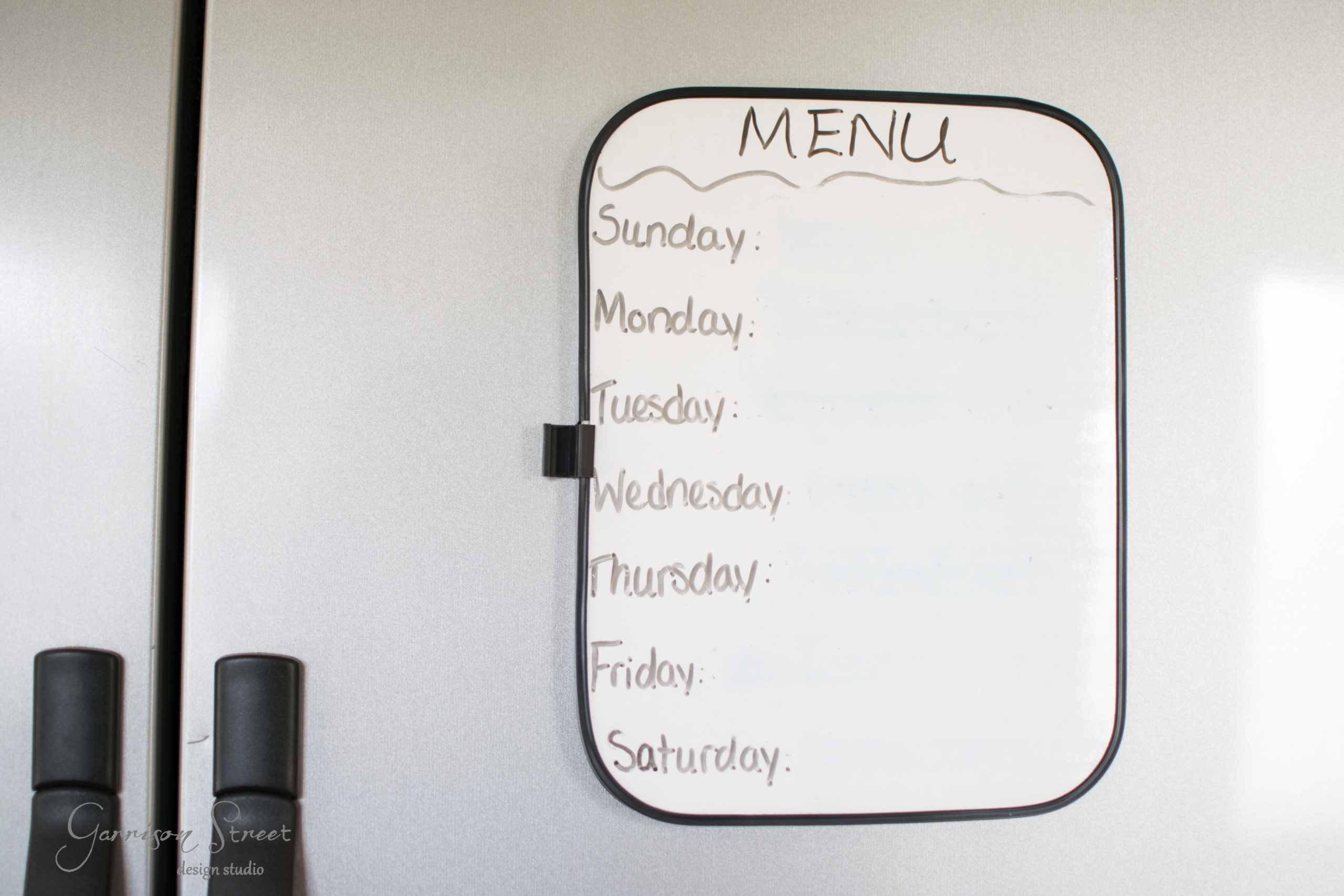 A whiteboard attached to a refrigerator lists days of the week with "MENU" written at the top. The list includes Sunday through Saturday, but no menu items are written beside the days. Two black handles of the refrigerator are visible.
